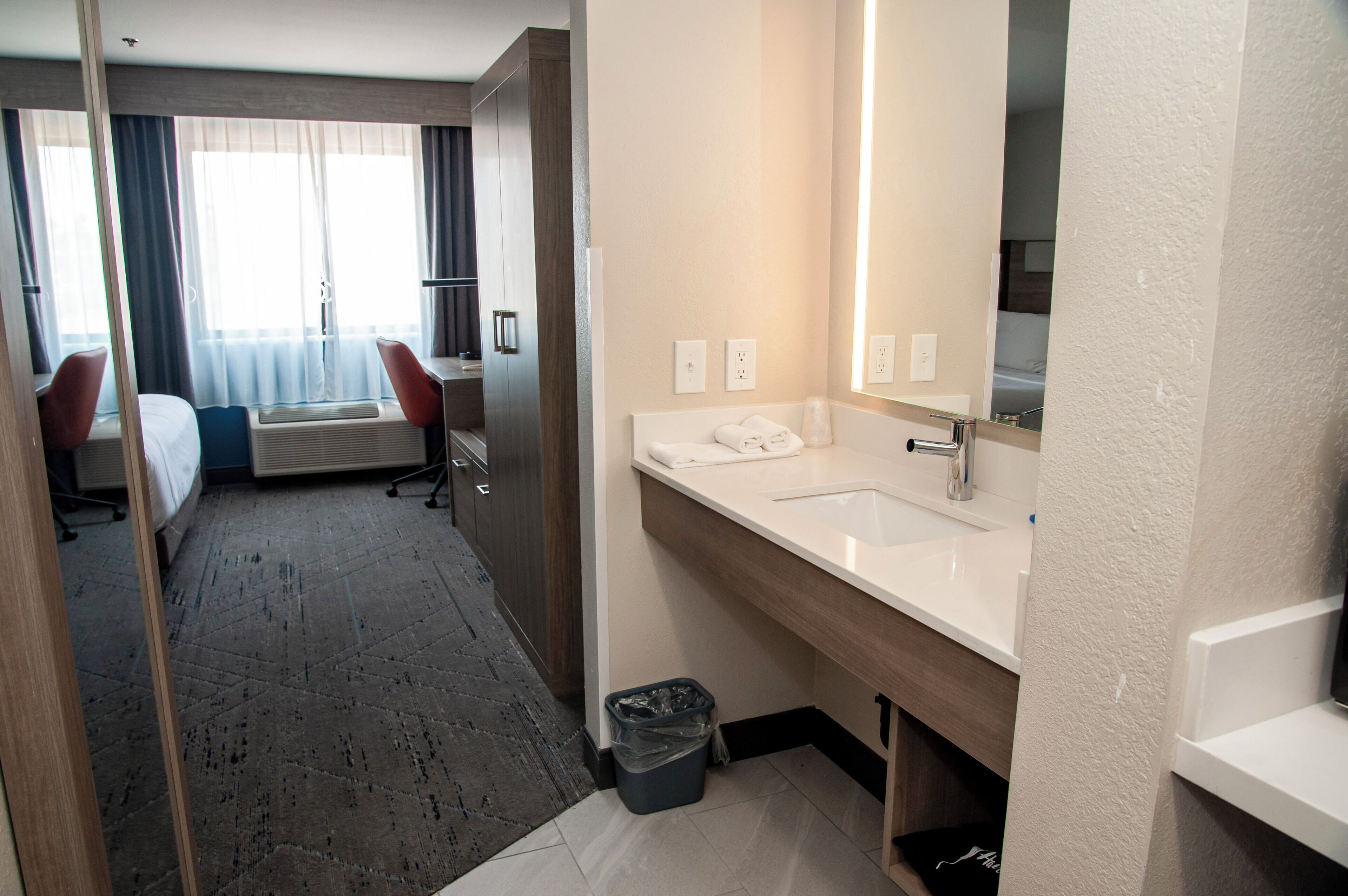 A hotel room with a bed, a desk, a chair, a large window with curtains, a bathroom with a sink and mirror, and a trash bin.