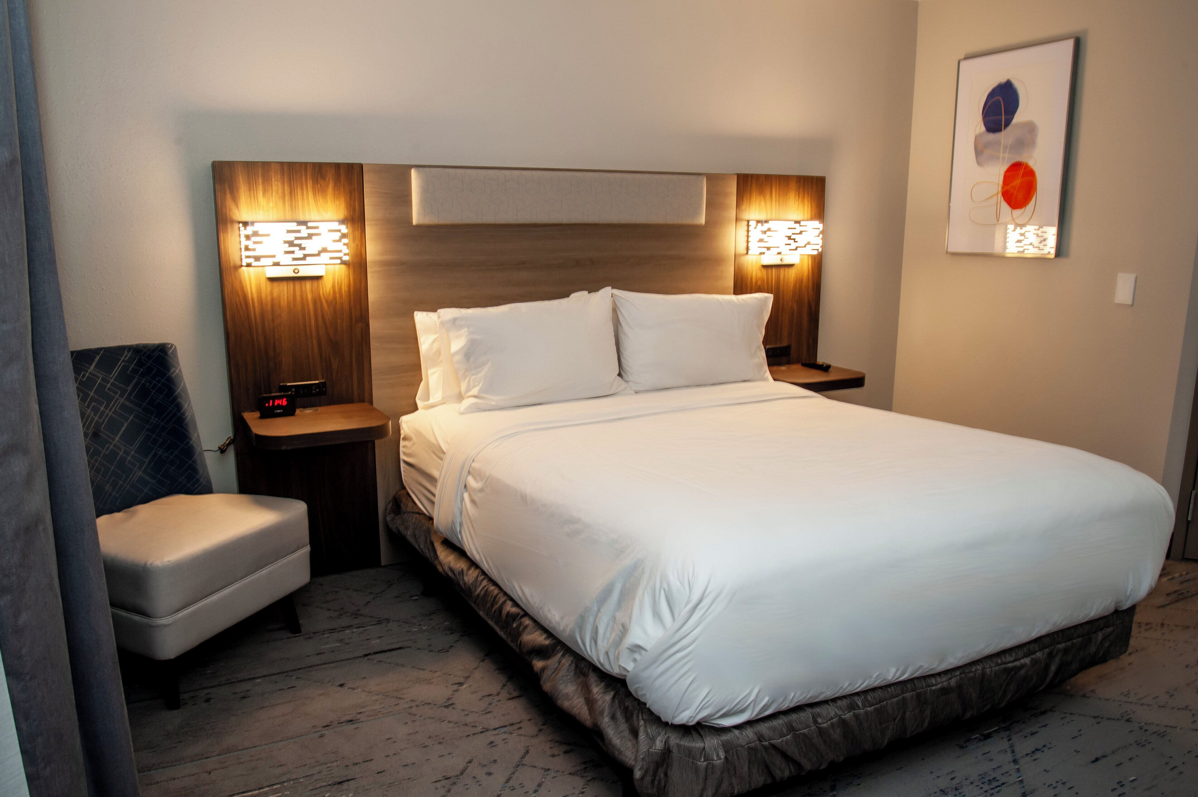A neatly made bed with white linens, two bedside tables with lamps, a chair, and a wall-mounted artwork.