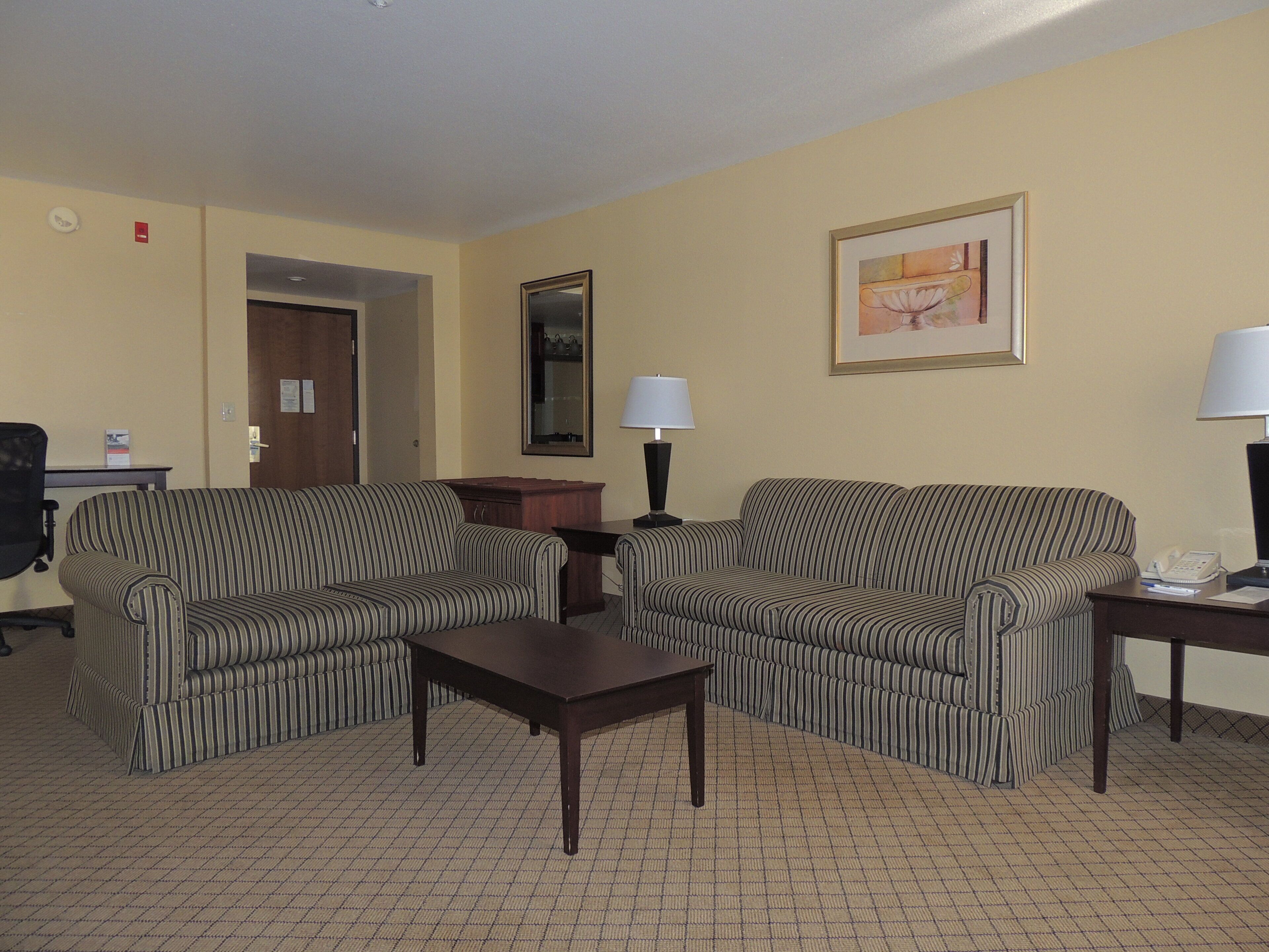 A hotel room with striped sofas, a small table, a lamp, a framed picture on the wall, and a desk with a telephone.