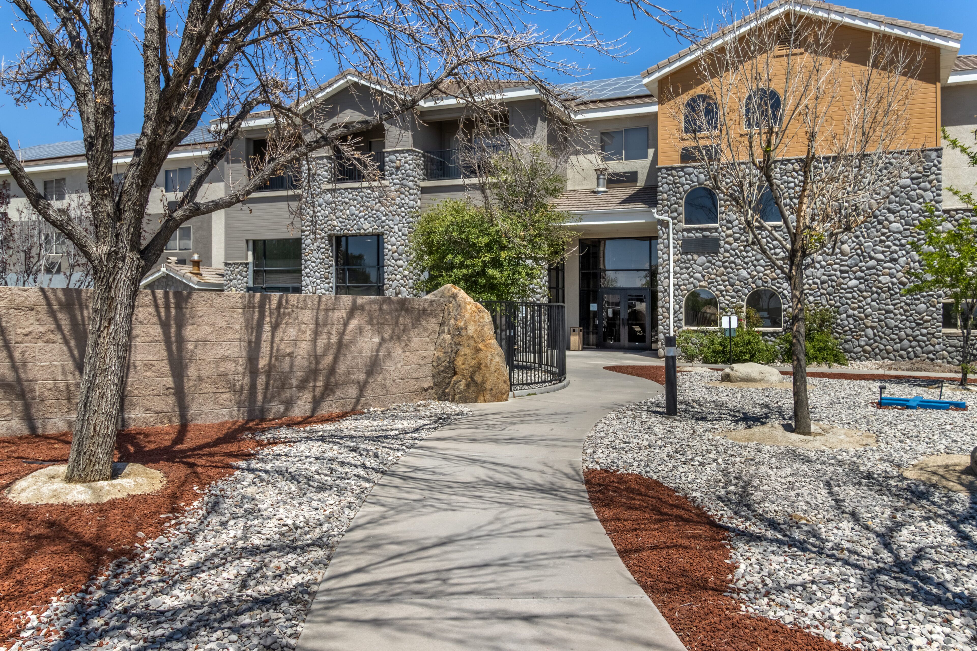 A modern building with a stone facade, a covered walkway, and a landscaped area with trees and gravel.