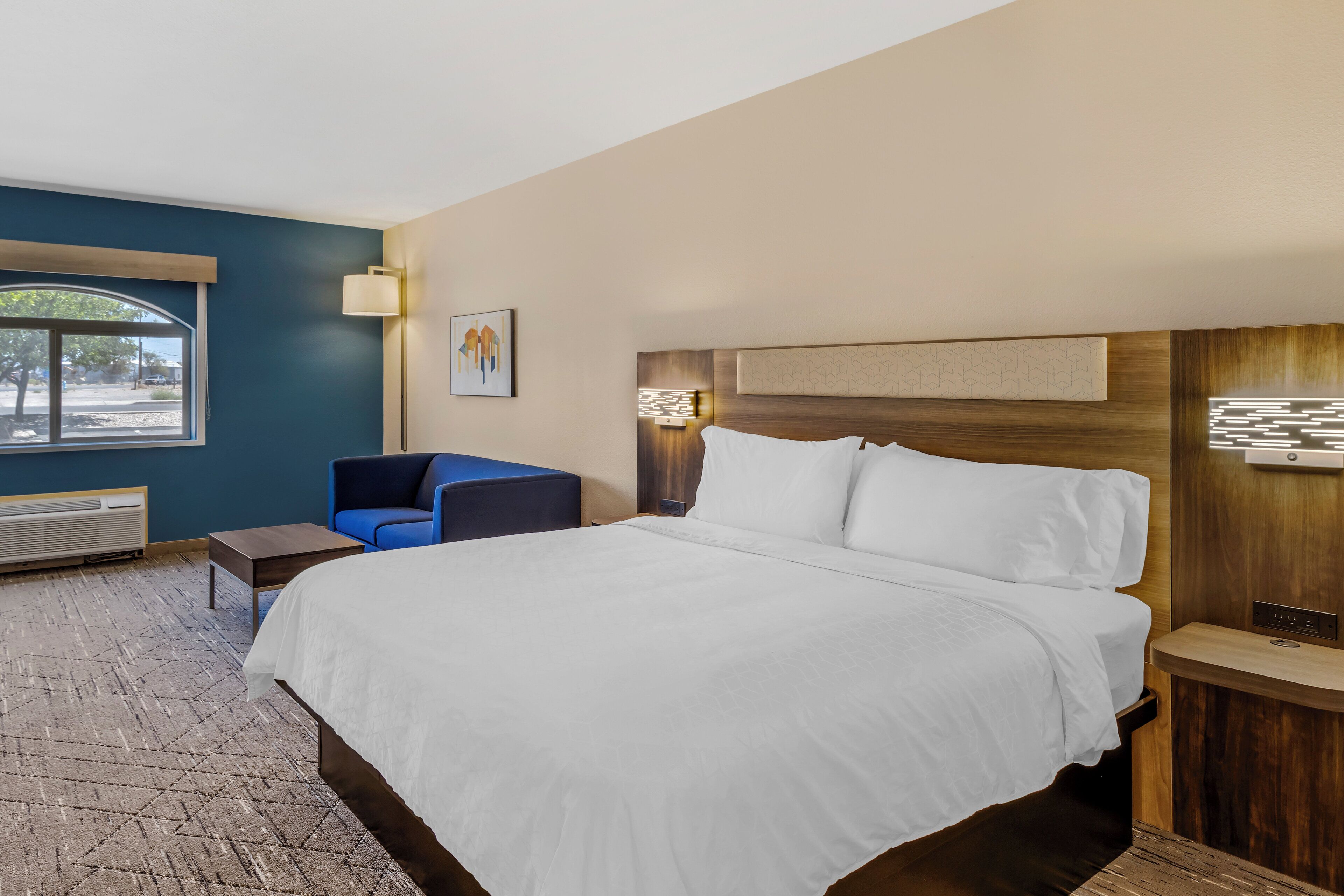 Habitación de hotel con una cama grande, un sillón azul, una mesita y una ventana con vistas al exterior.