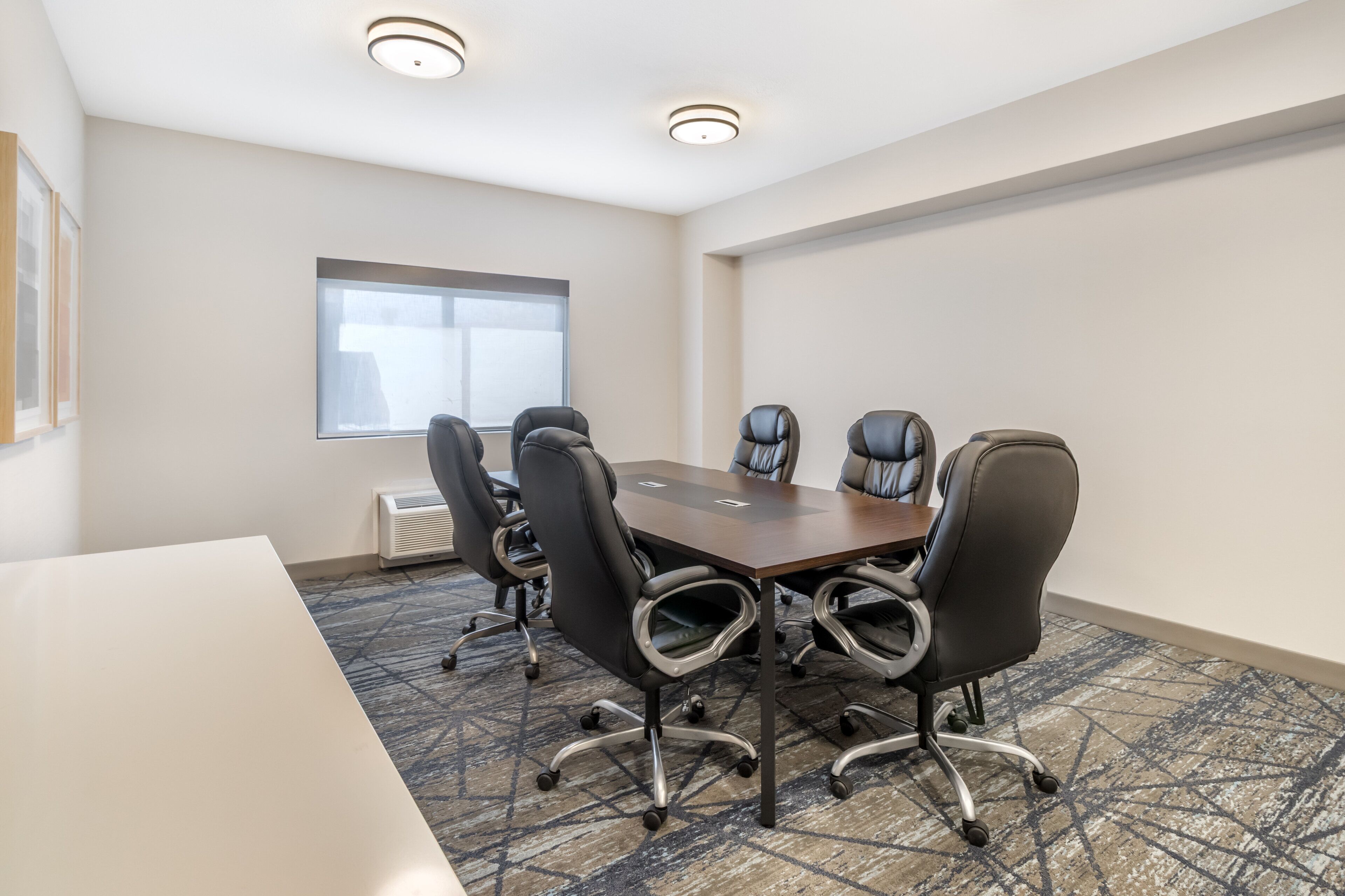A conference room with a rectangular table, six black office chairs, a window with blinds, and a ceiling light fixture.