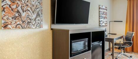 A hotel room with a flat-screen TV mounted above a minibar, a desk with a chair, and a patterned carpet.