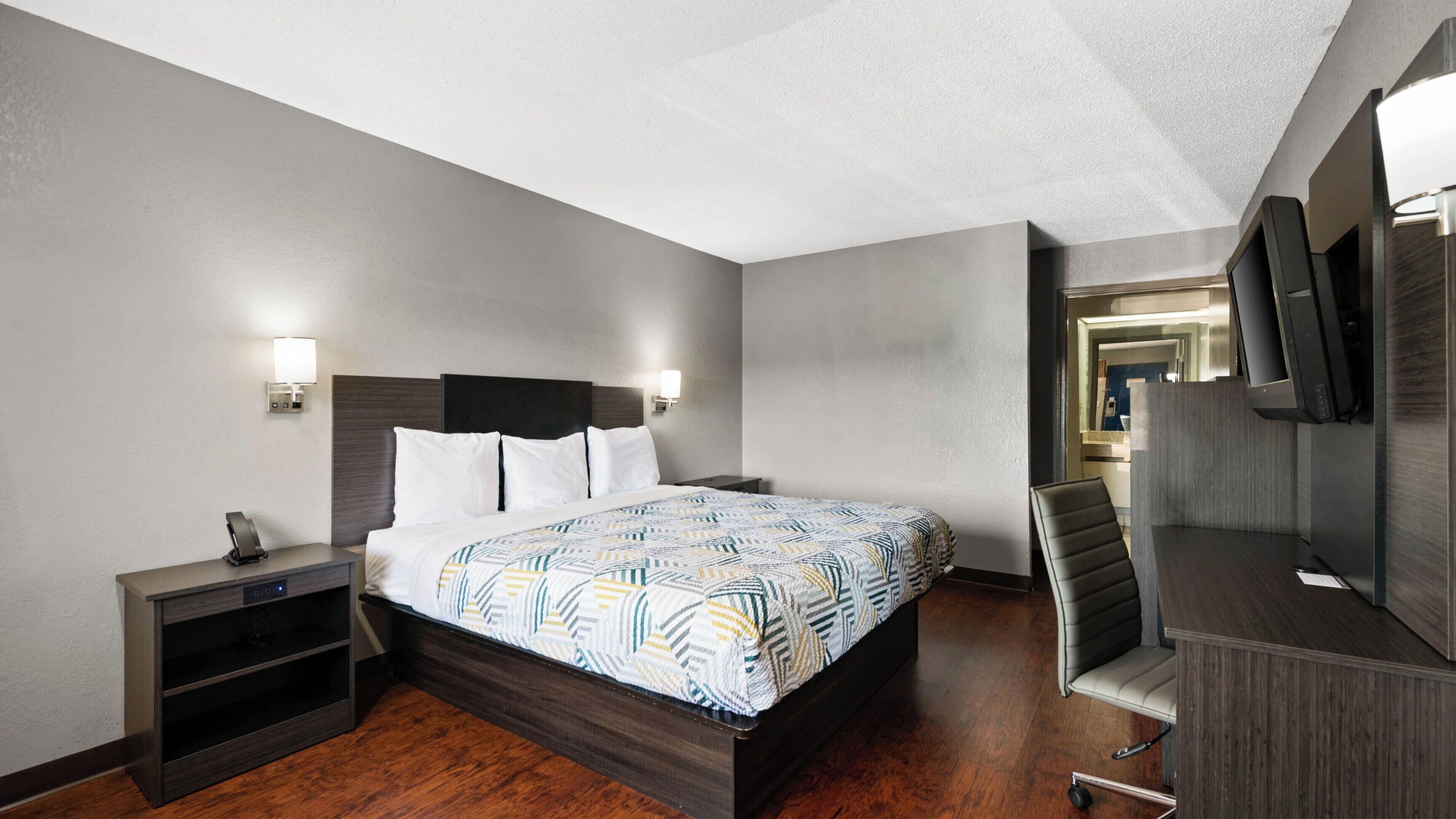 Een hotelkamer met een bed, nachtkastje, lamp, bureau, stoel en een aan de muur bevestigde televisie.