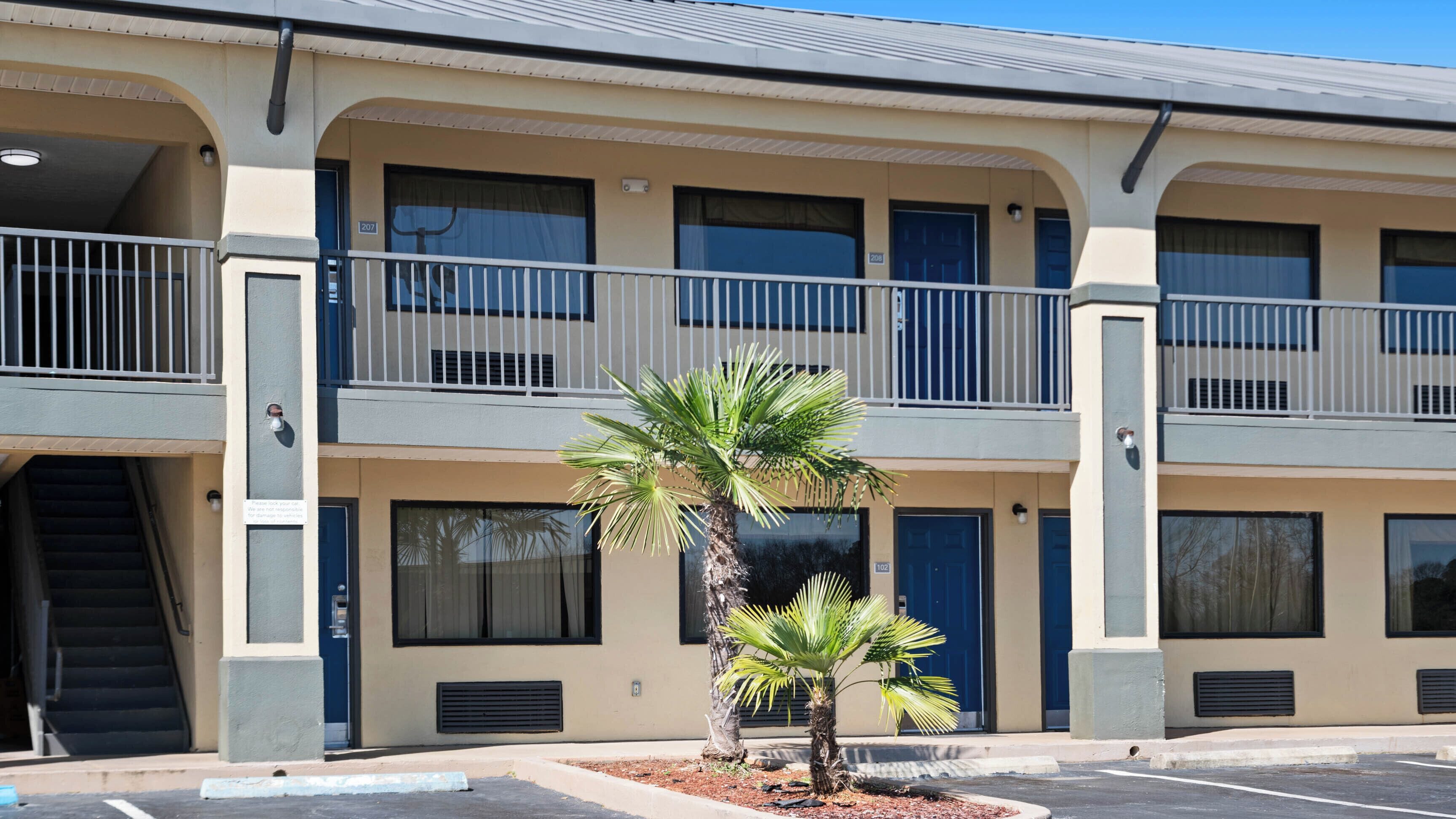 A multi-story motel with balconies, a parking lot, and a palm tree in the foreground.