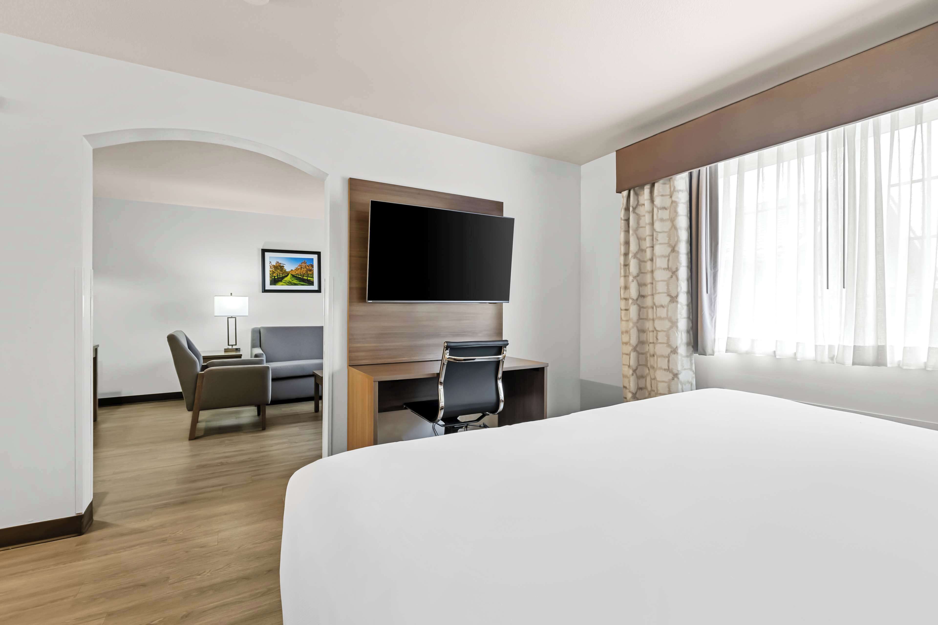 Habitación de hotel con una cama grande, un escritorio con silla, un televisor de pantalla plana en la pared y una ventana con cortinas translúcidas.