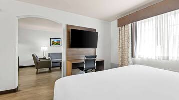 Habitación de hotel con una cama grande, un escritorio con silla, un televisor de pantalla plana en la pared y una ventana con cortinas translúcidas.