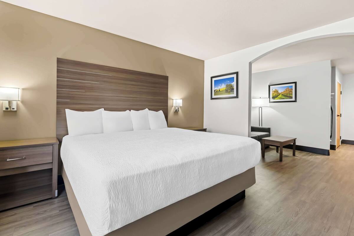 Une chambre d’hôtel moderne avec un grand lit, une tête de lit en bois, deux tables de chevet avec des lampes, une petite table de chevet et deux tableaux encadrés au mur.