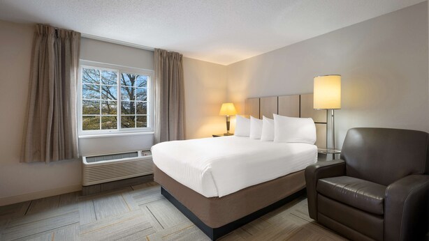 Een hotelkamer met een groot bed, twee bedlampjes, een bruine leren fauteuil en een raam met gordijnen.