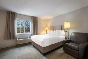 Una habitación de hotel con una cama grande, dos lámparas de noche, un sillón de cuero marrón y una ventana con cortinas.