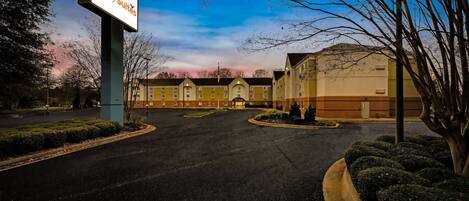A hotel entrance with a large sign, a parking lot, and a building with a yellow facade.