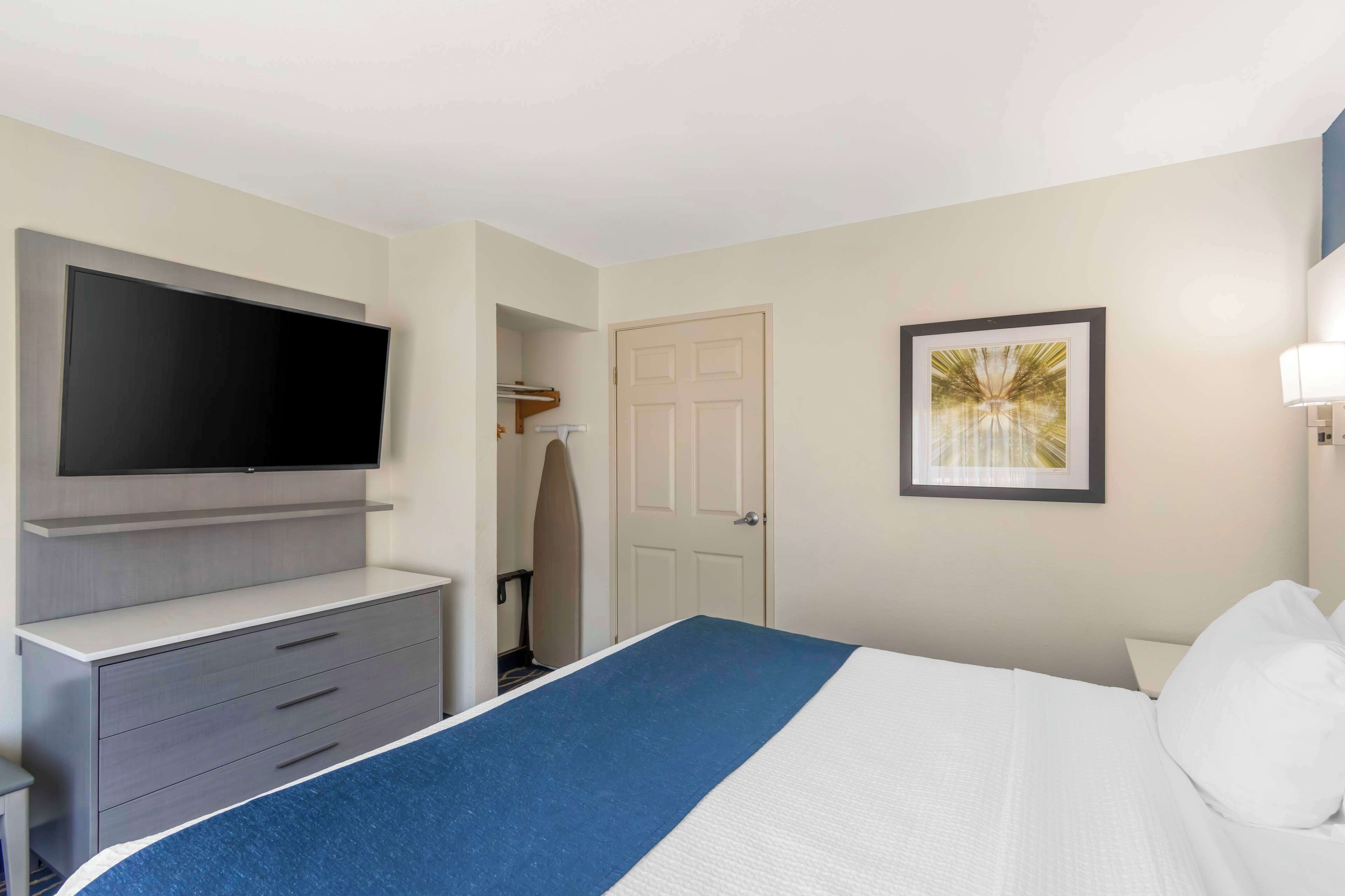 A hotel room with a bed, a flat-screen TV mounted on a dresser, a door, and a framed picture on the wall.