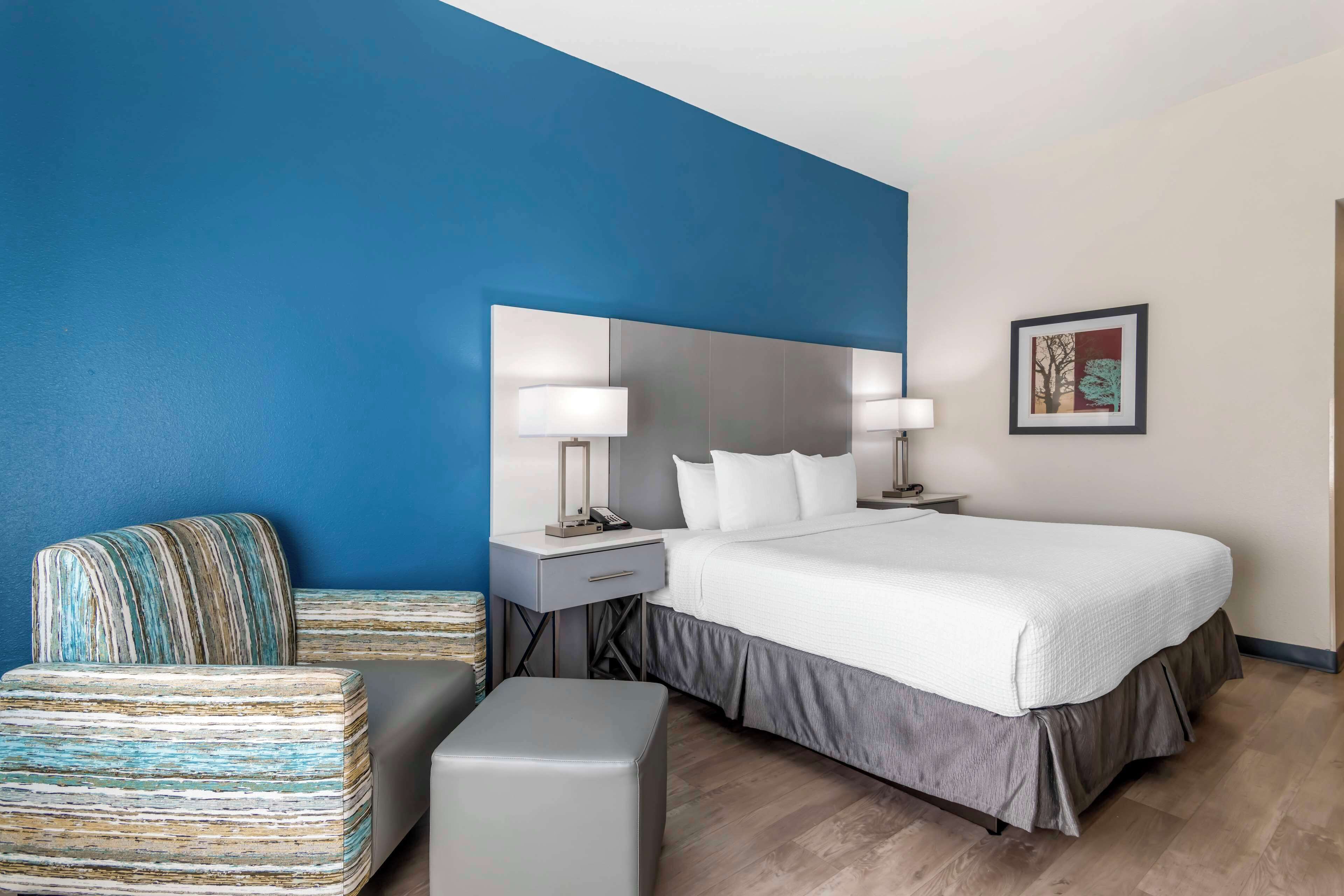 Una habitación de hotel moderna con una cama grande, una mesita de noche con lámpara, una silla e imagen enmarcada en la pared.