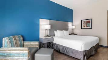 Una habitación de hotel moderna con una cama grande, una mesita de noche con lámpara, una silla e imagen enmarcada en la pared.