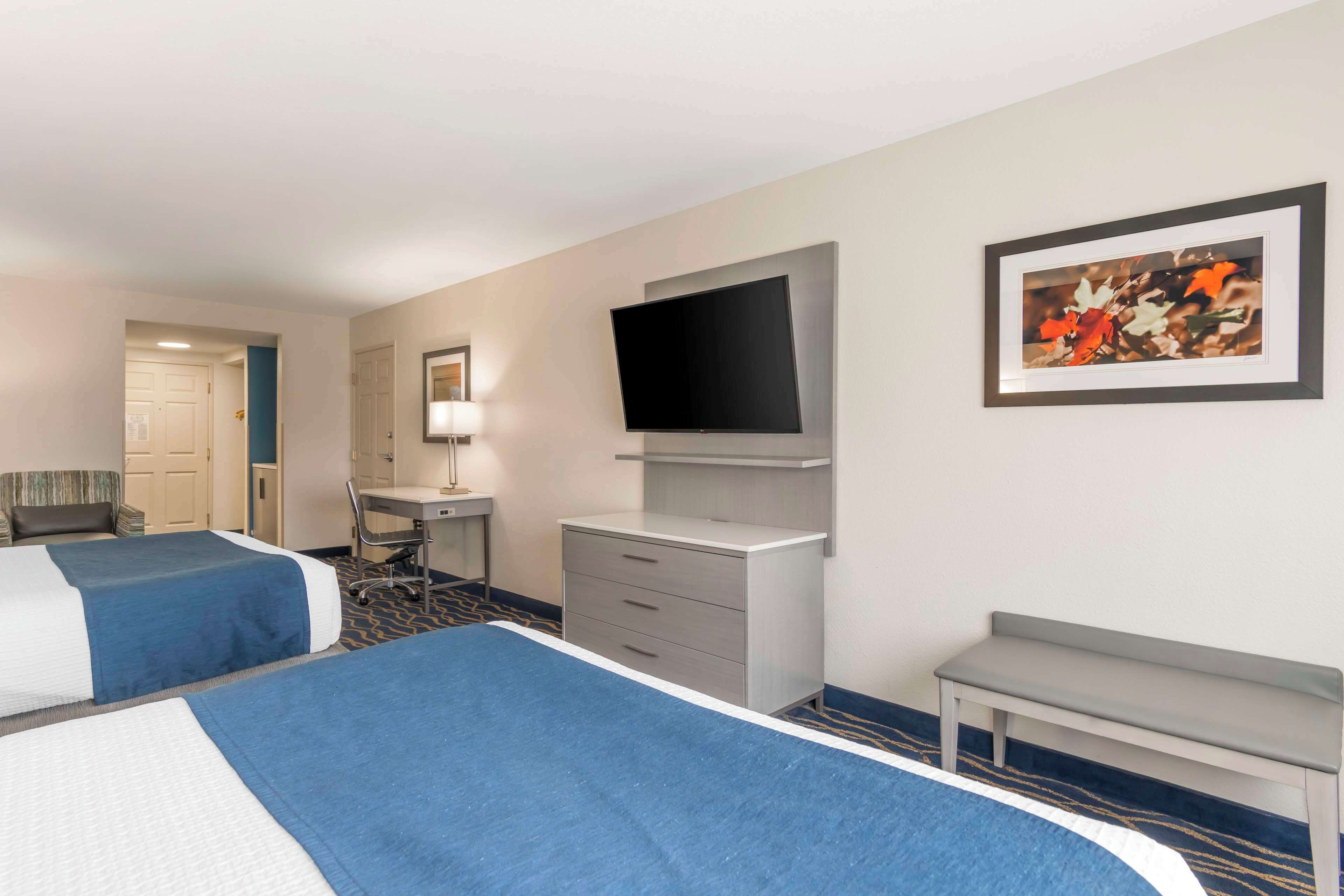 A hotel room with two beds, a desk, a chair, a television, and a bench.