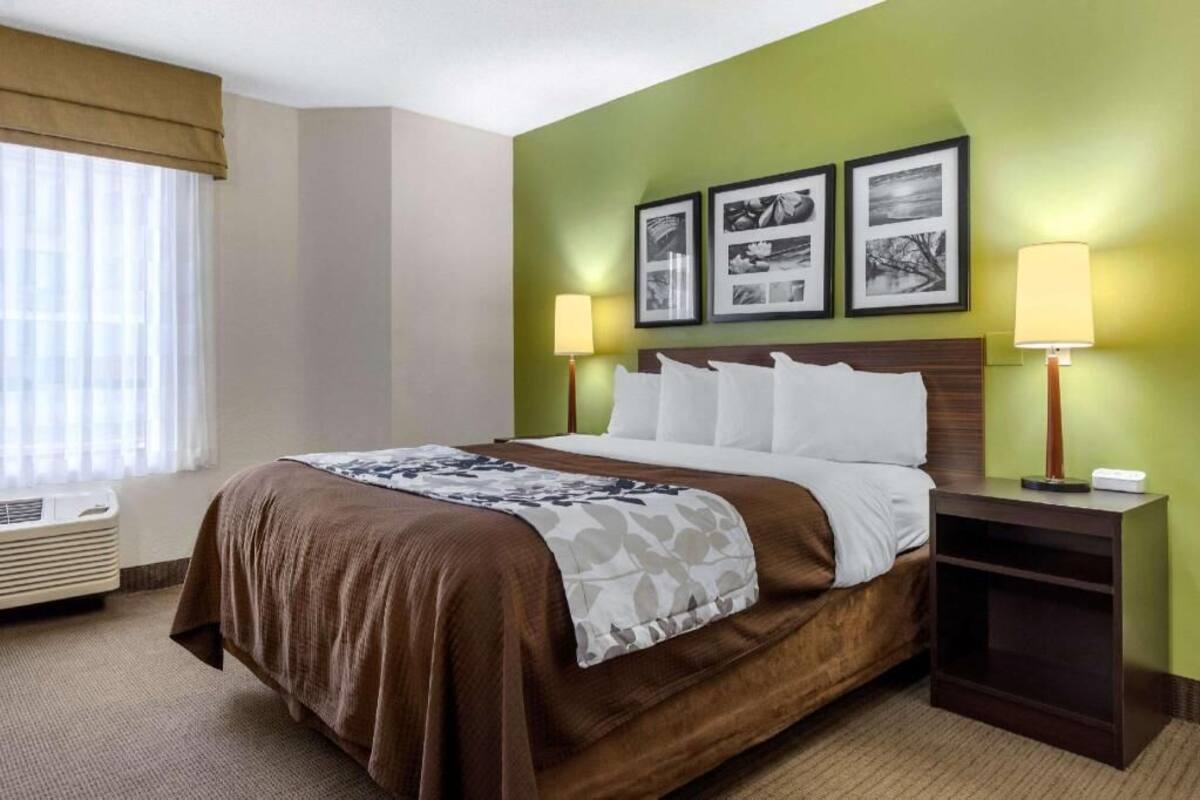 A hotel room with a bed, two bedside lamps, a window with blinds, and a wall with three framed pictures.