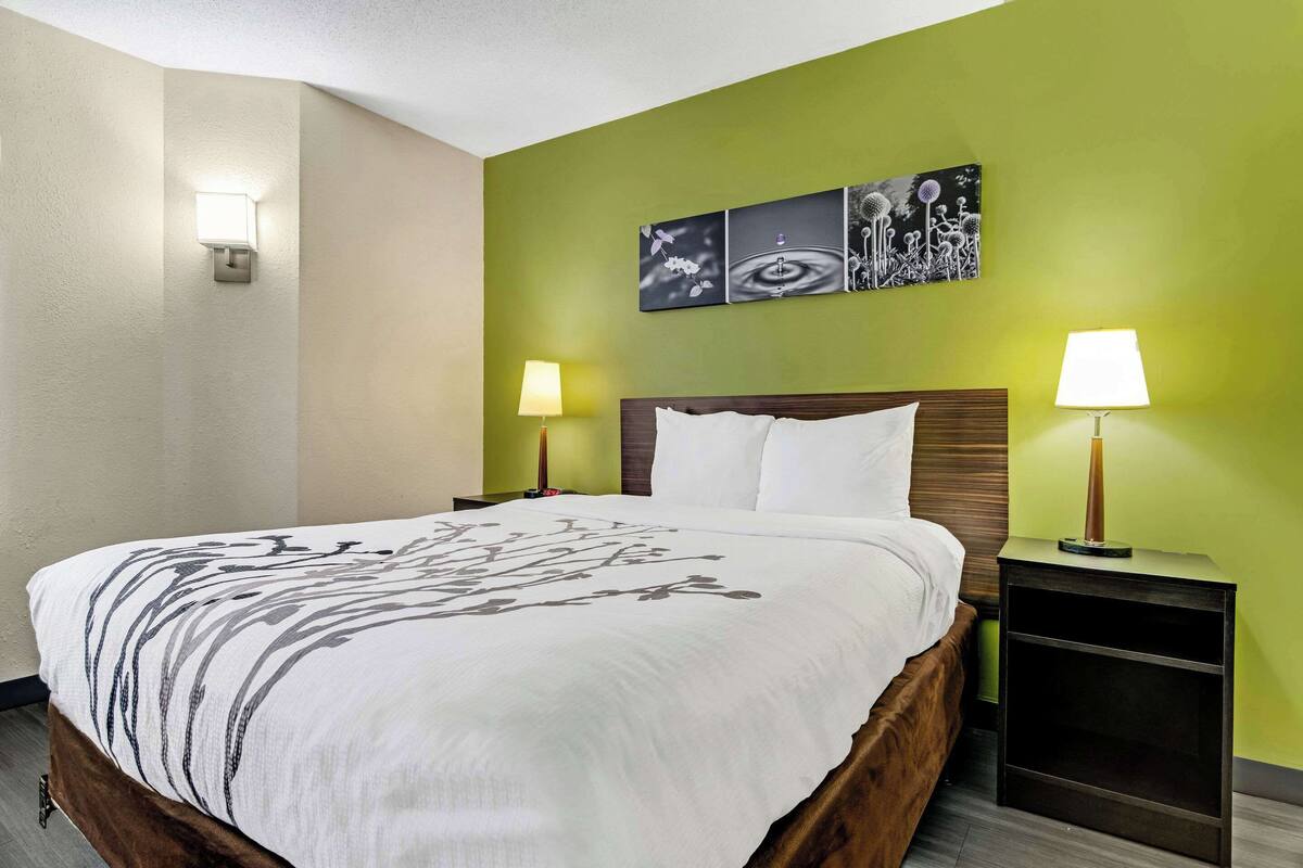 A neatly made bed with a white and gray patterned bedspread, two bedside lamps, a nightstand, and a wall with three black and white artwork pieces.