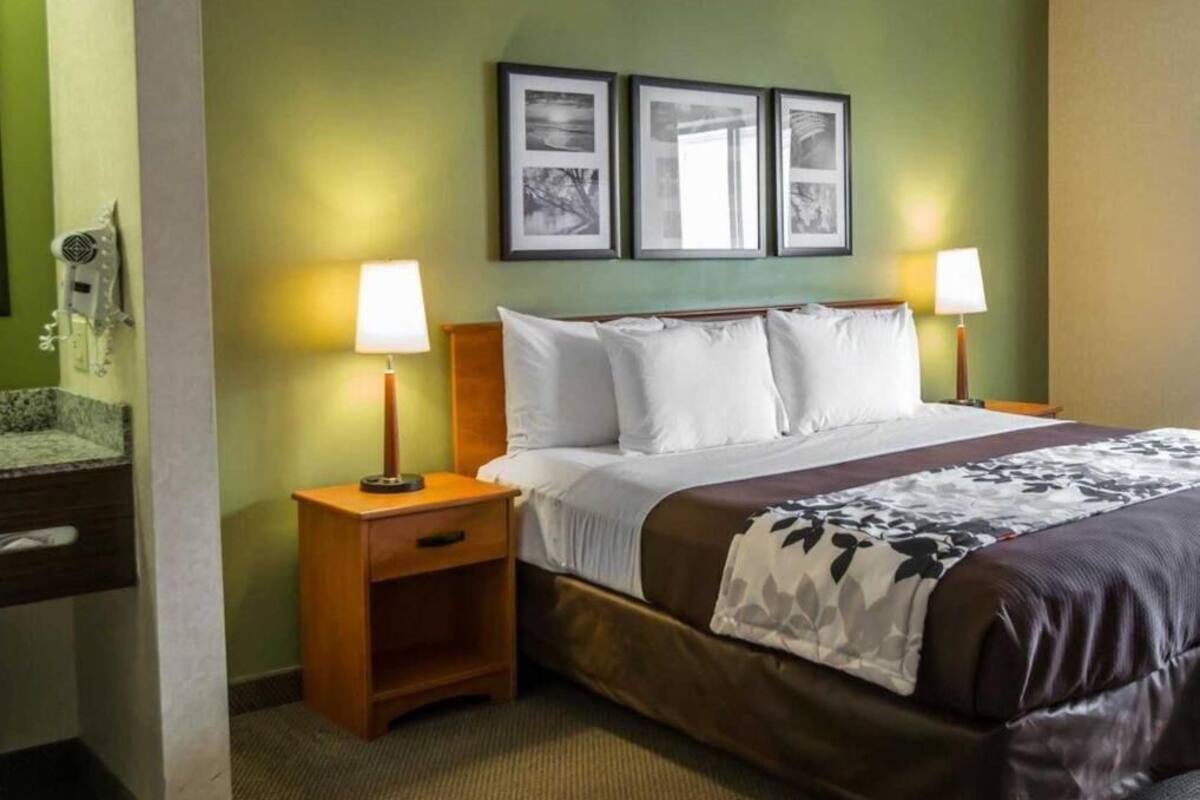 A hotel room with a bed, two bedside tables with lamps, a wooden nightstand, a wall-mounted shelf, and framed pictures above the bed.