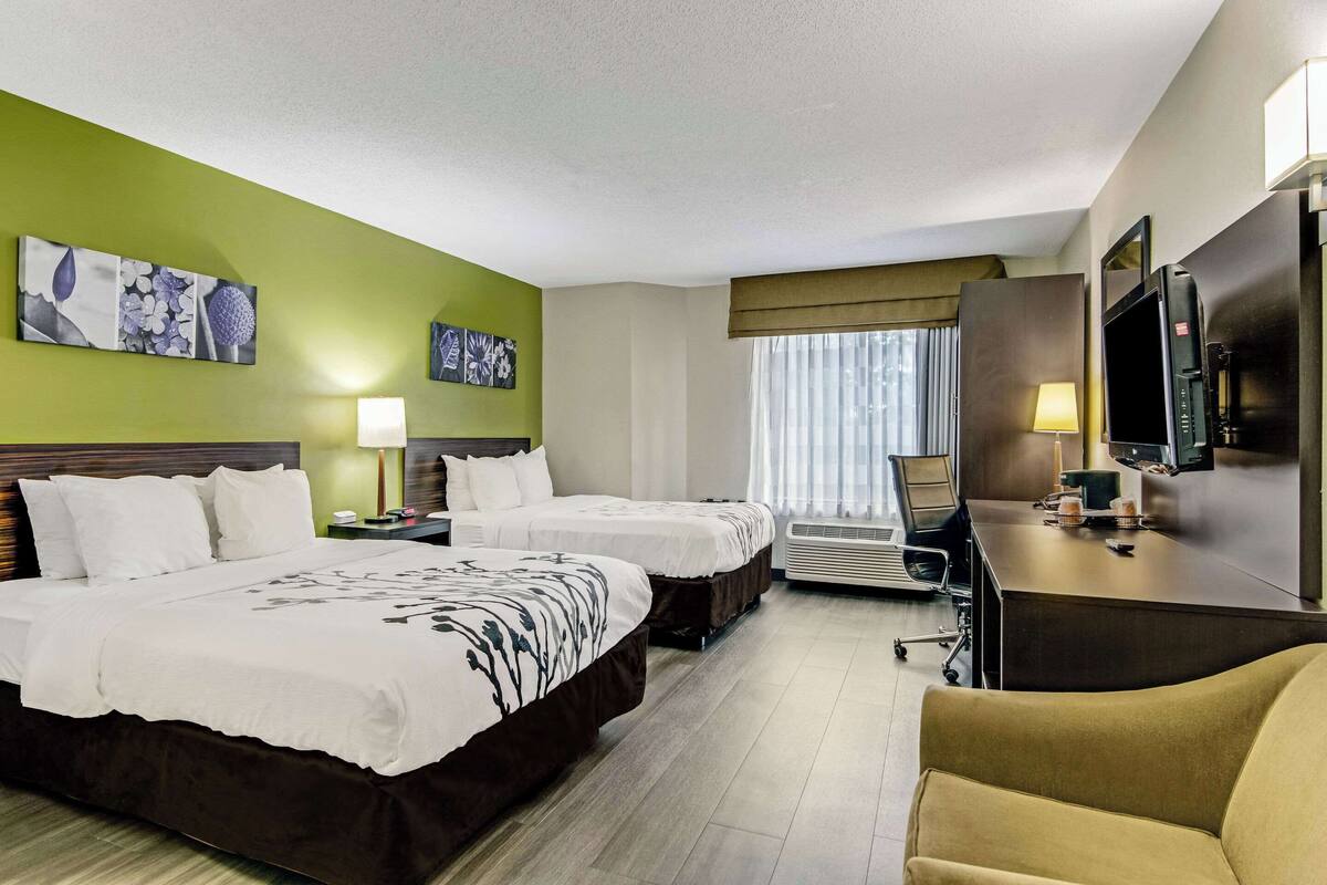 A hotel room with two beds, a desk with a TV, a chair, a desk lamp, and artwork on the walls.