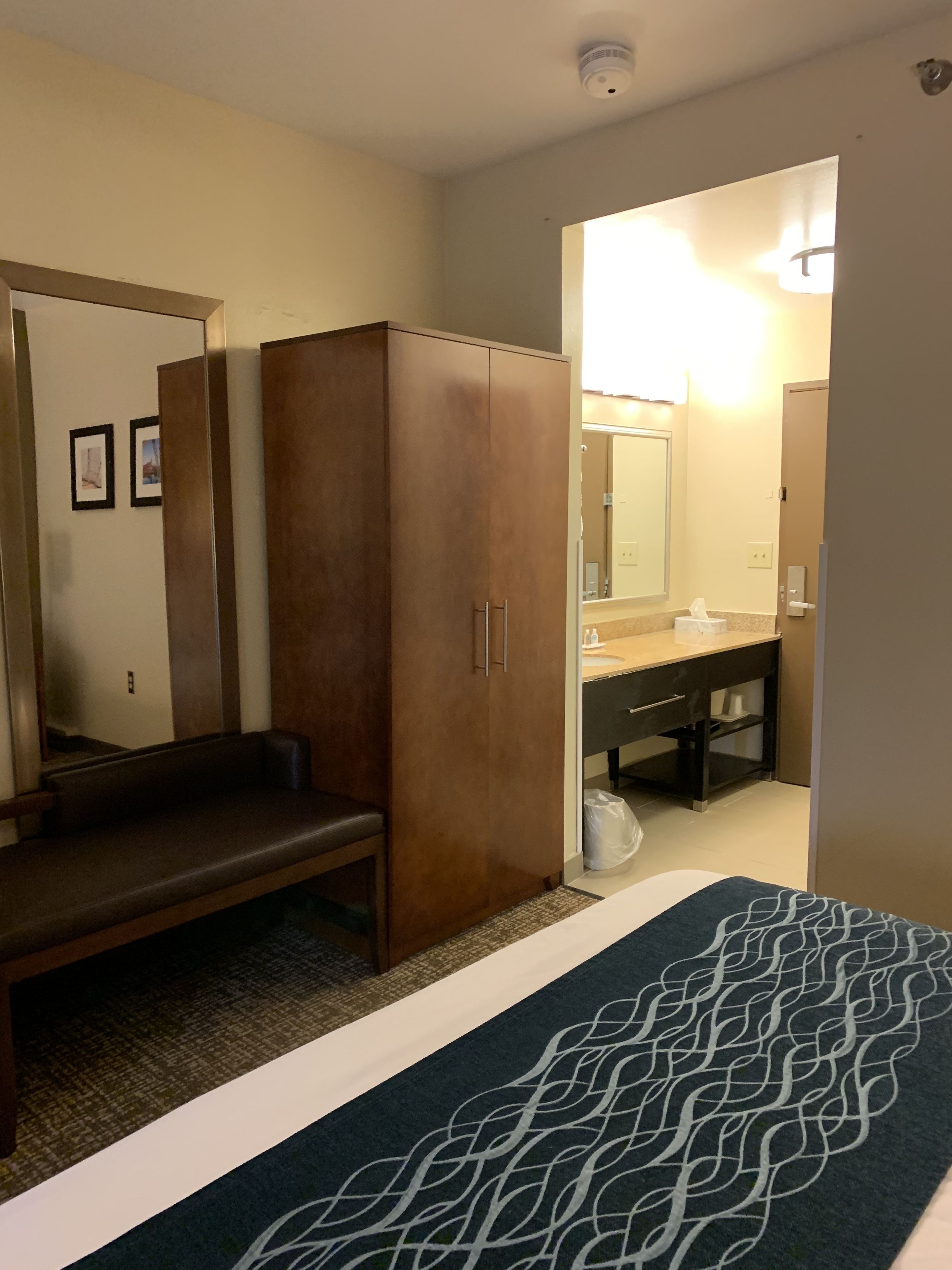 Una habitación de hotel con cama, armario de madera, banco, baño con espejo y cesto de basura.