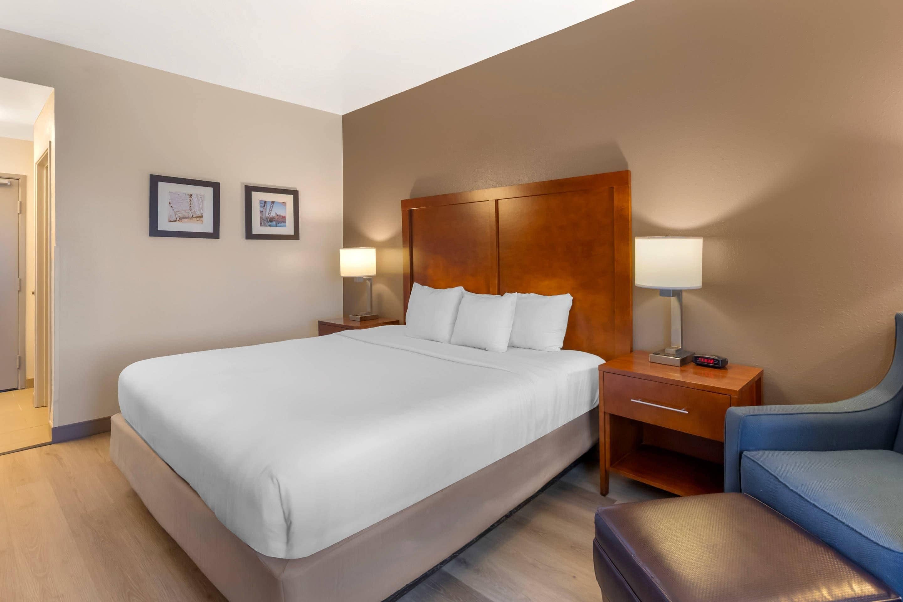 Una habitación de hotel con una cama grande, mesitas de noche con lámparas, un sillón y dos cuadros en la pared.