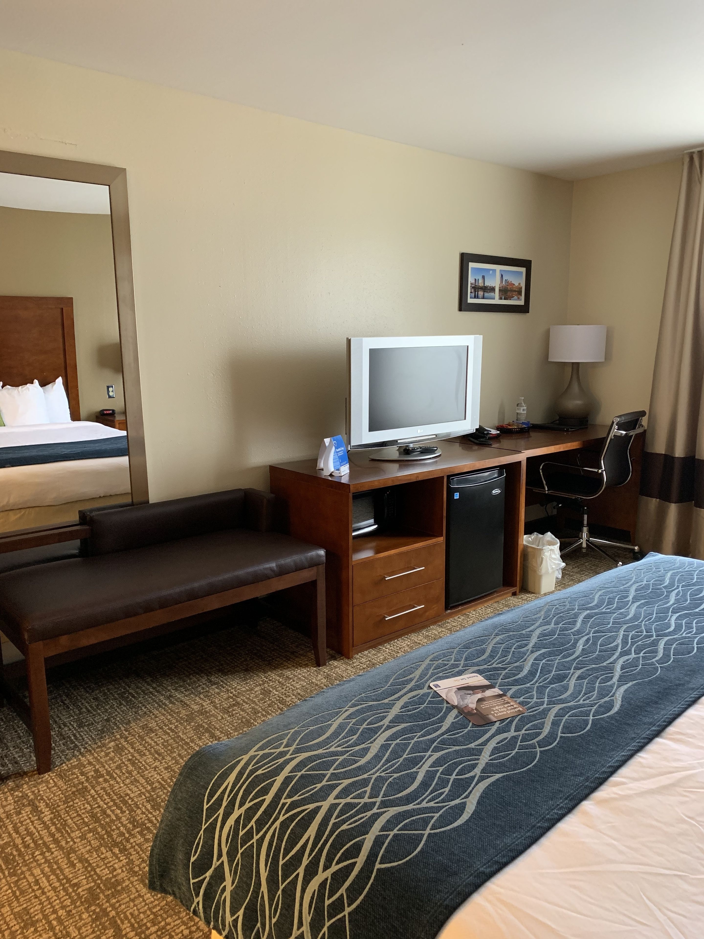 A hotel room with a bed, a desk with a television, a chair, a lamp, and a small trash bin.
