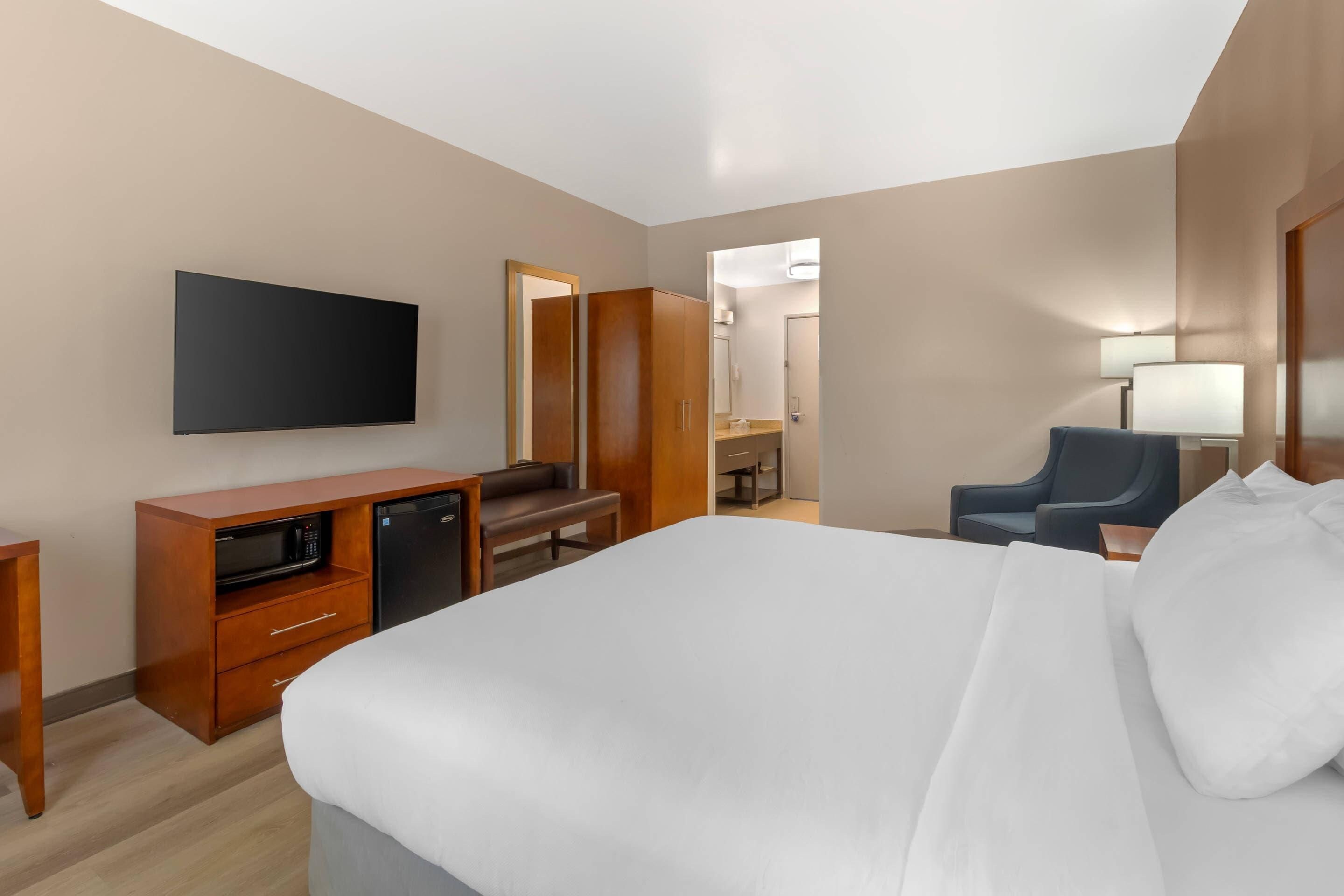 A hotel room with a bed, a desk with a computer, a TV, a chair, and a bathroom visible through an open door.
