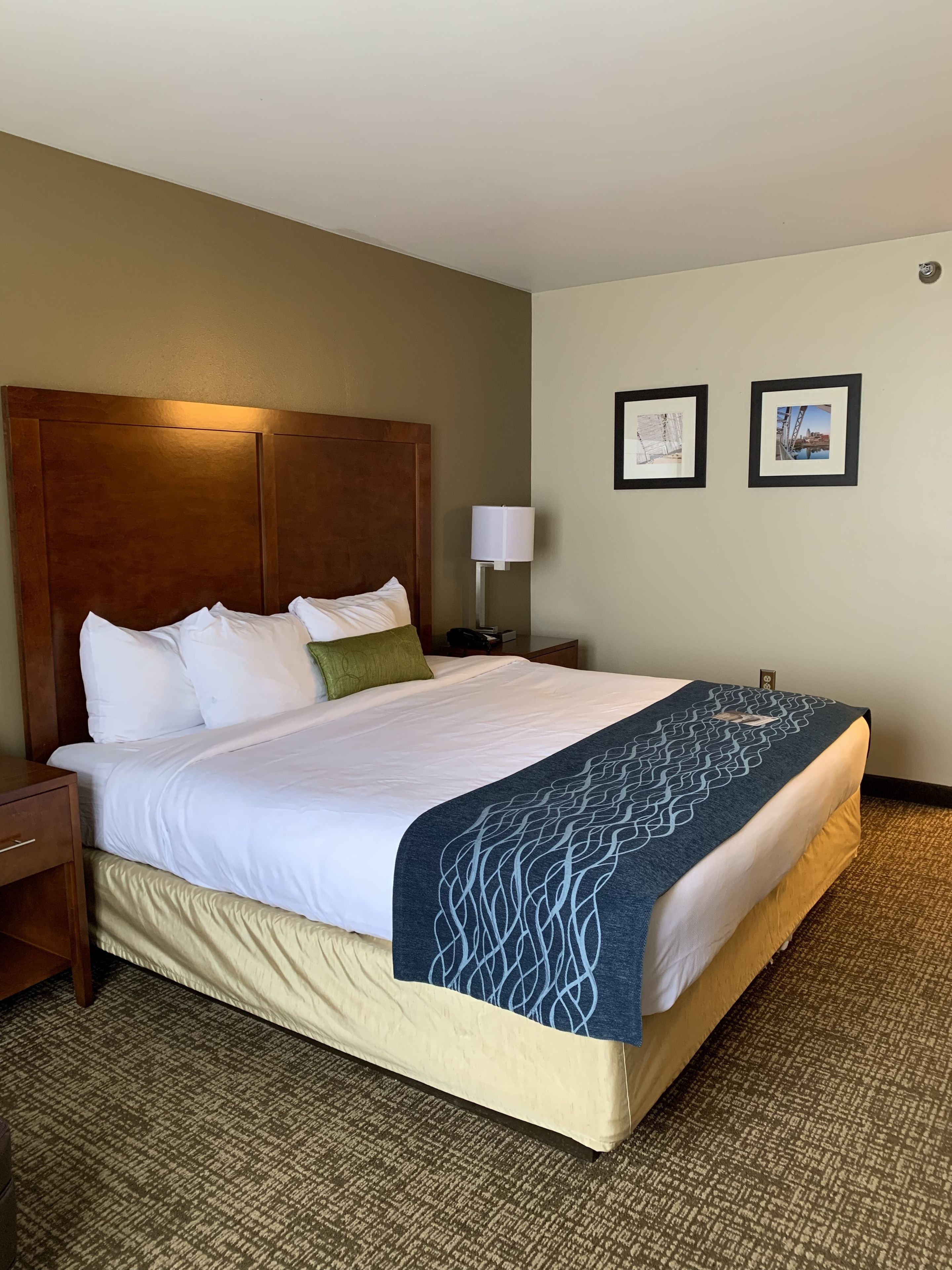 A hotel room with a large bed, a wooden headboard, a nightstand, a lamp, and two framed pictures on the wall.