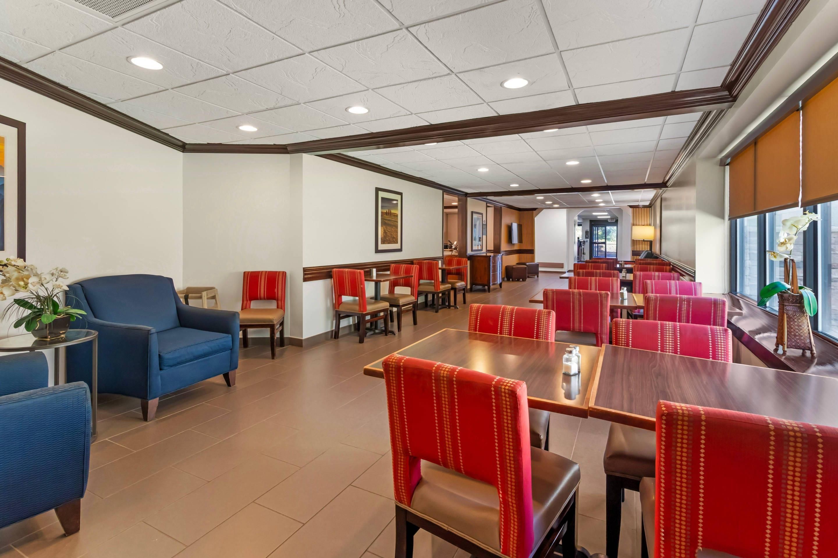 Un comedor de hotel con asientos tipo banca tapizados en rojo y mesas, un sofá azul y un amplio ventanal con luz natural.