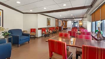 Uma sala de jantar de hotel com bancos estofados vermelhos e mesas, um sofá azul e uma grande janela com luz natural.