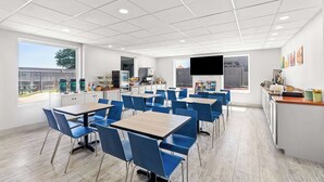 A modern dining area with multiple tables and chairs, a kitchen area with appliances, and large windows offering views of the outside.