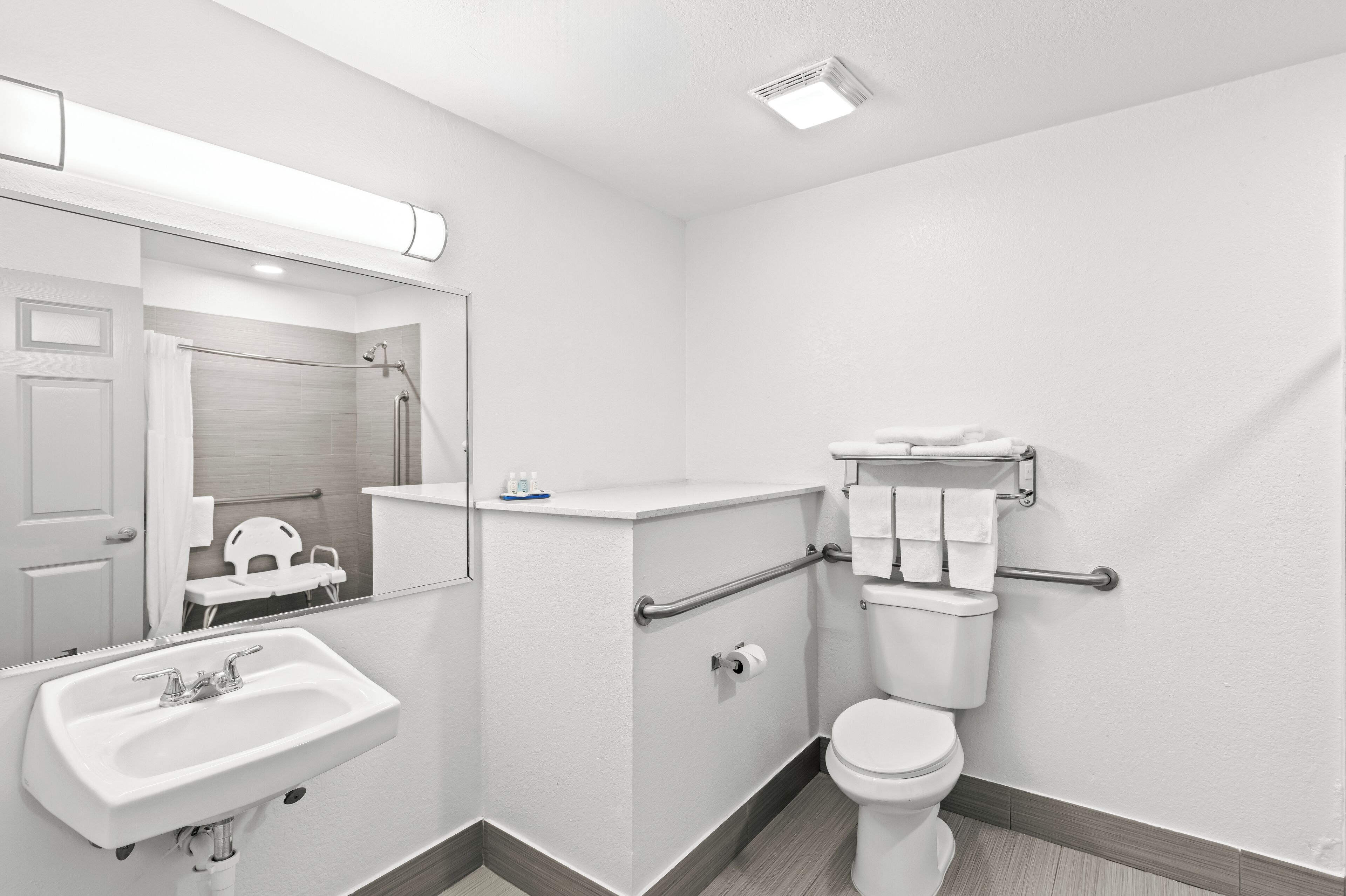 A modern bathroom with a white sink, toilet, and mirror. It includes grab bars and a towel rack.