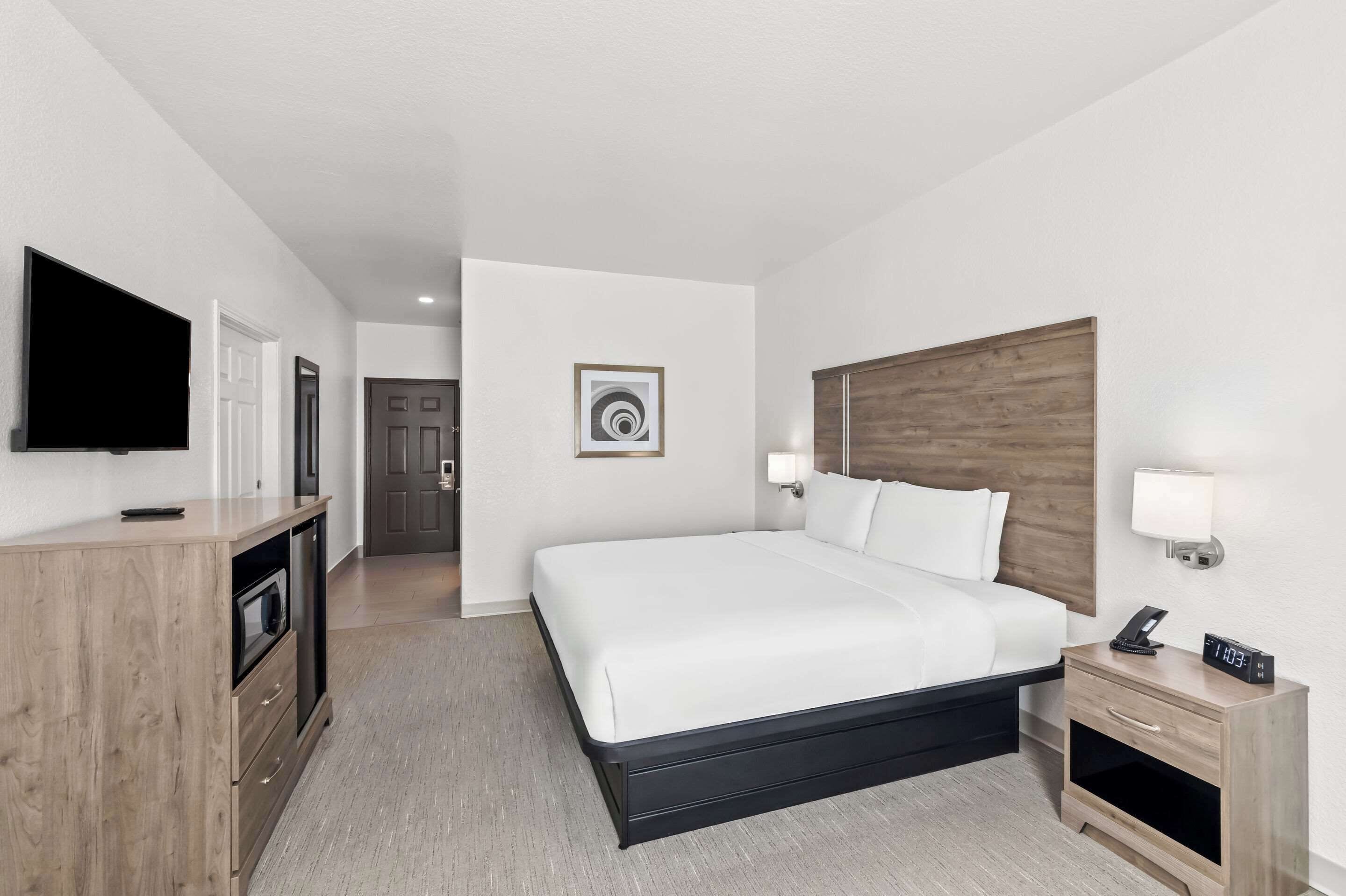 Una camera d'albergo con un letto grande, una TV, una scrivania con una lampada e un comodino con una sveglia.