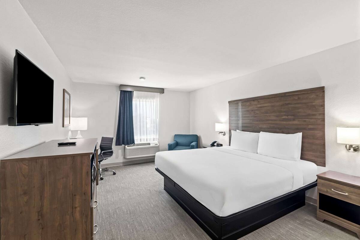 A hotel room with a large bed, a desk with a chair, a TV mounted on the wall, and a window with curtains.