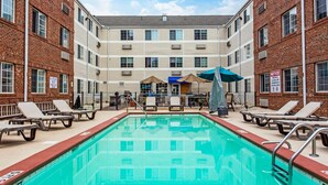 Una piscina con sillas de descanso y sombrillas, rodeada de edificios de ladrillo con varias ventanas y unidades de aire acondicionado.