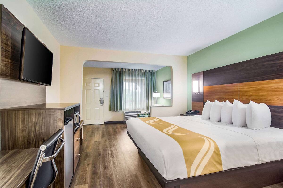 A hotel room with a bed, a desk, a television, and a bathroom visible through an archway.