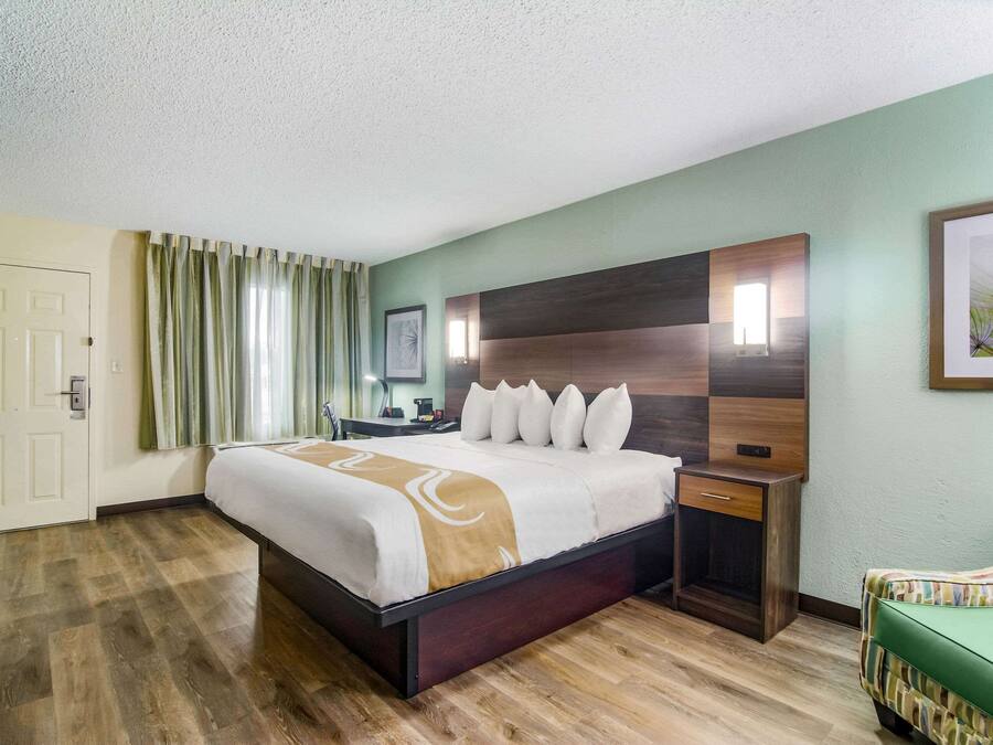Una habitación de hotel con una cama grande, un cabecero de madera, una mesita de noche, un sillón verde y una puerta.