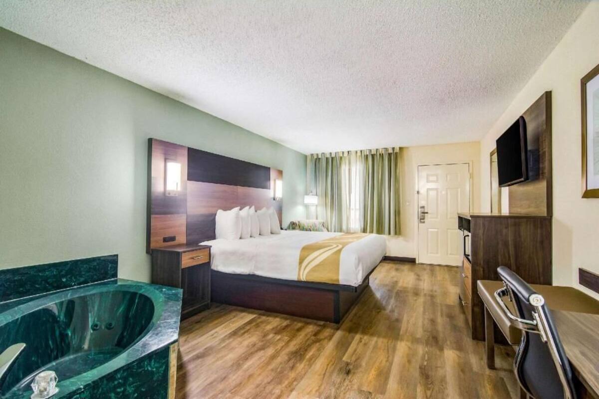 A hotel room with a bed, a TV, a desk, a chair, a bathroom with a bathtub, and a door.
