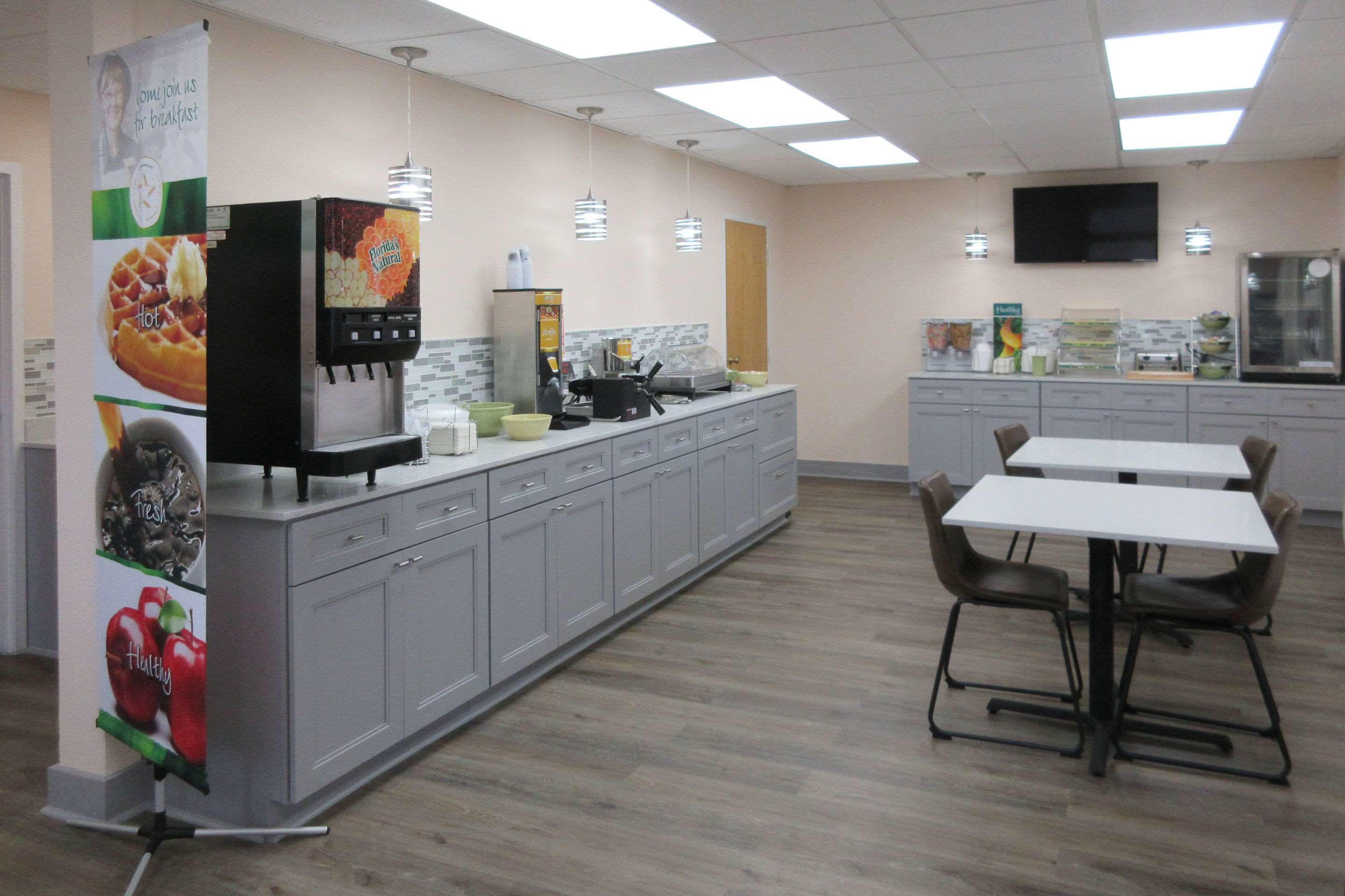 A modern kitchen area with a long counter, a coffee machine, and a microwave. There are two tables with chairs, a television mounted on the wall, and a window with blinds.