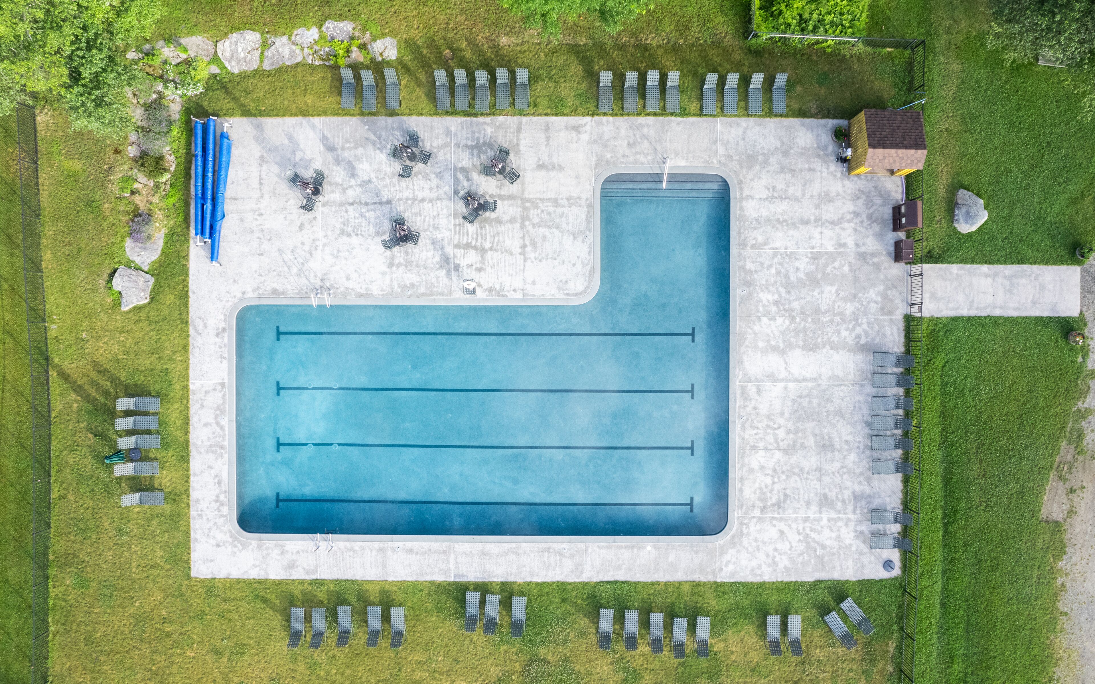 Eine Vogelperspektive auf ein Schwimmbecken mit mehreren Bahnen, umgeben von Liegestühlen und einer überdachten Sitzgelegenheit.