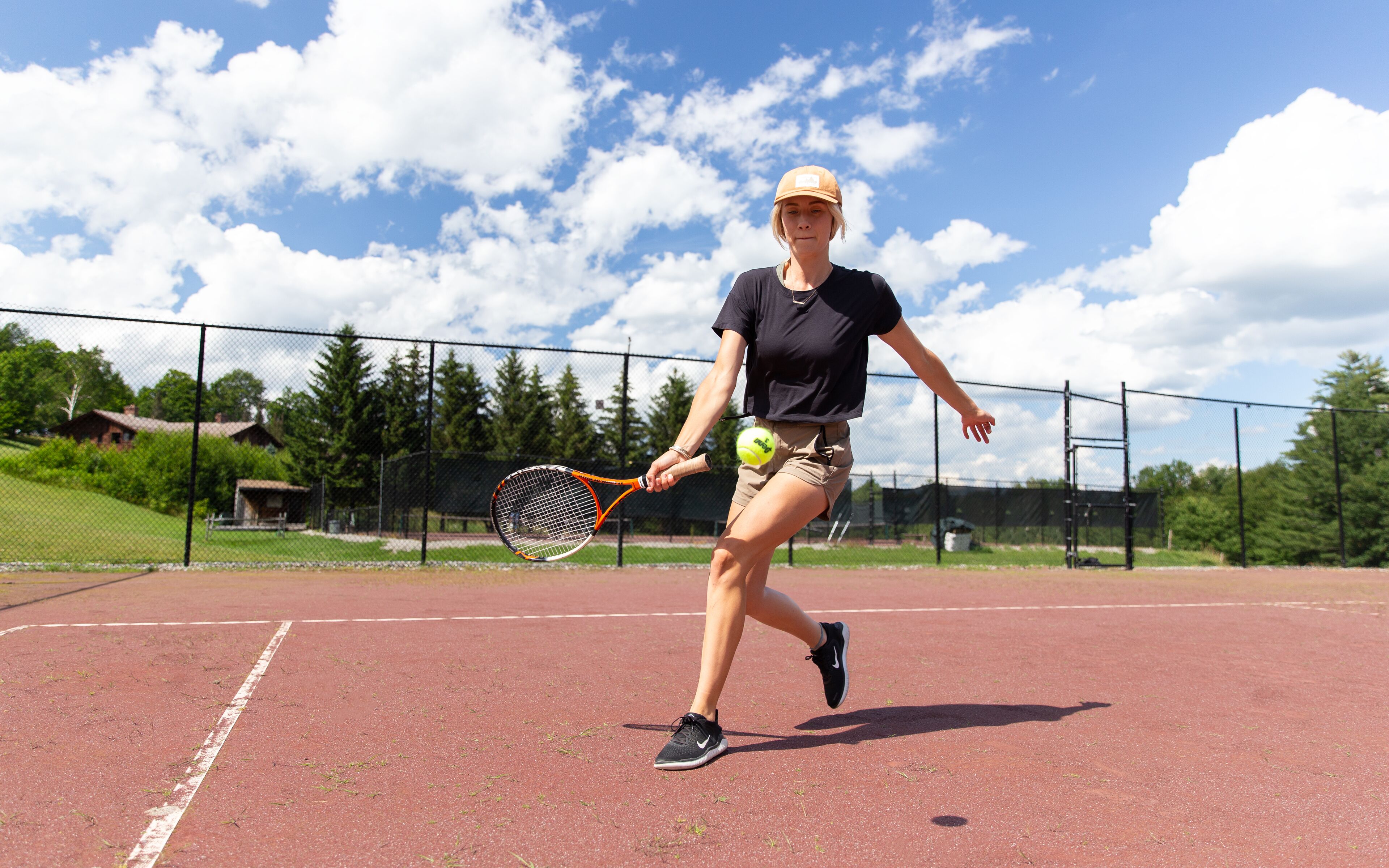 Un court de tennis avec un joueur en plein mouvement, une clôture et des arbres en arrière-plan.