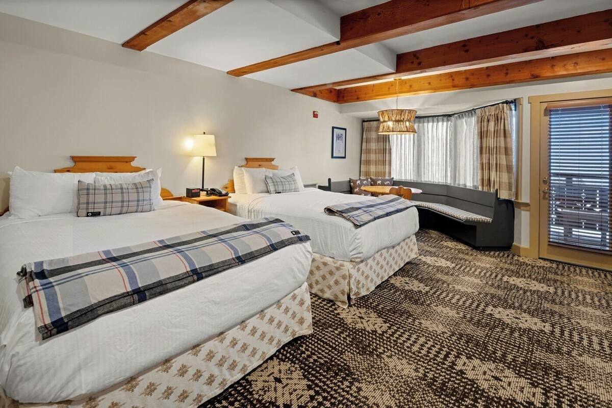 Una habitación de hotel con dos camas, un ventanal con cortinas, una alfombra estampada y vigas de madera en el techo.