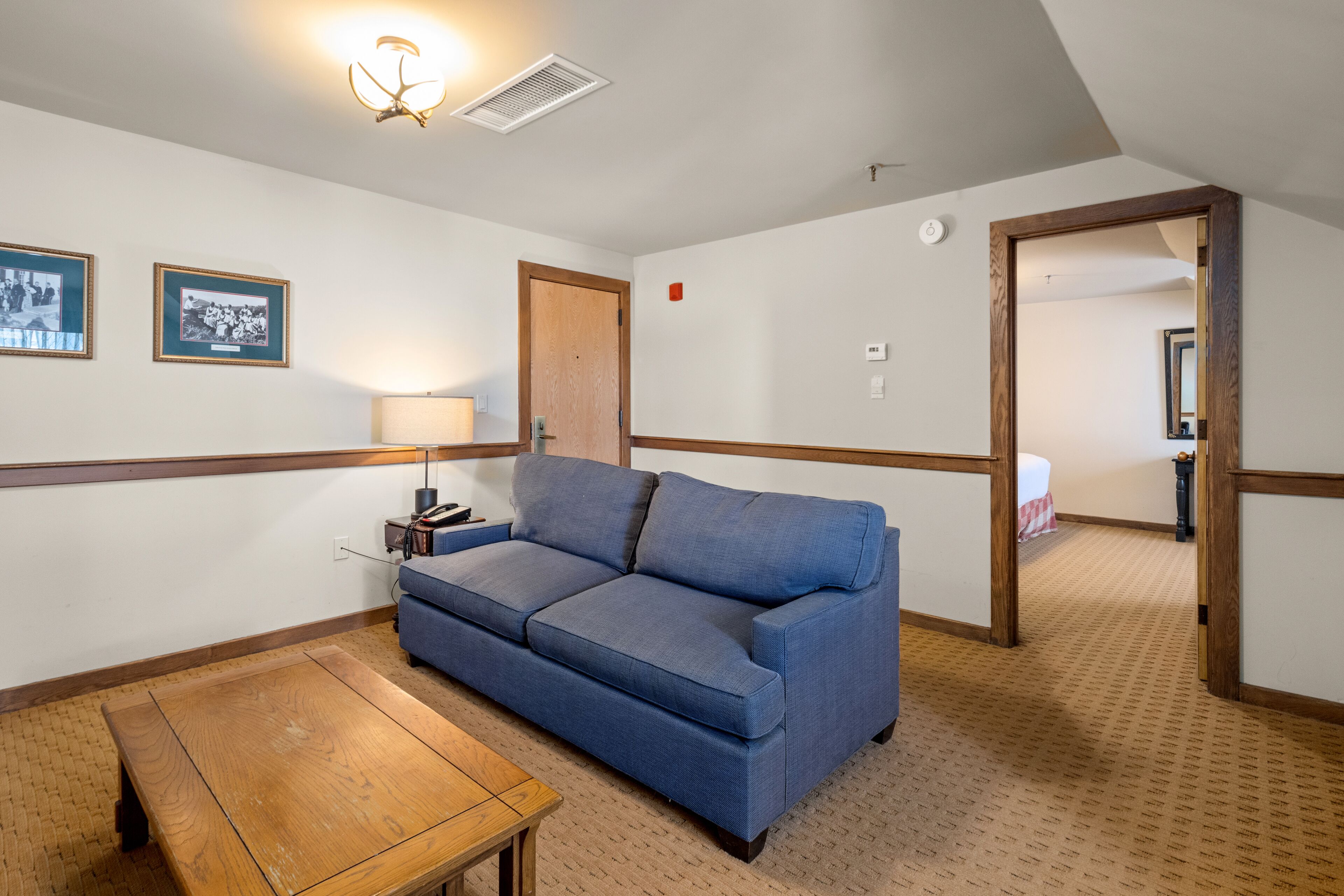Una habitación con un sofá azul, una mesa de madera, cuadros enmarcados en la pared y una puerta que da a otra habitación.