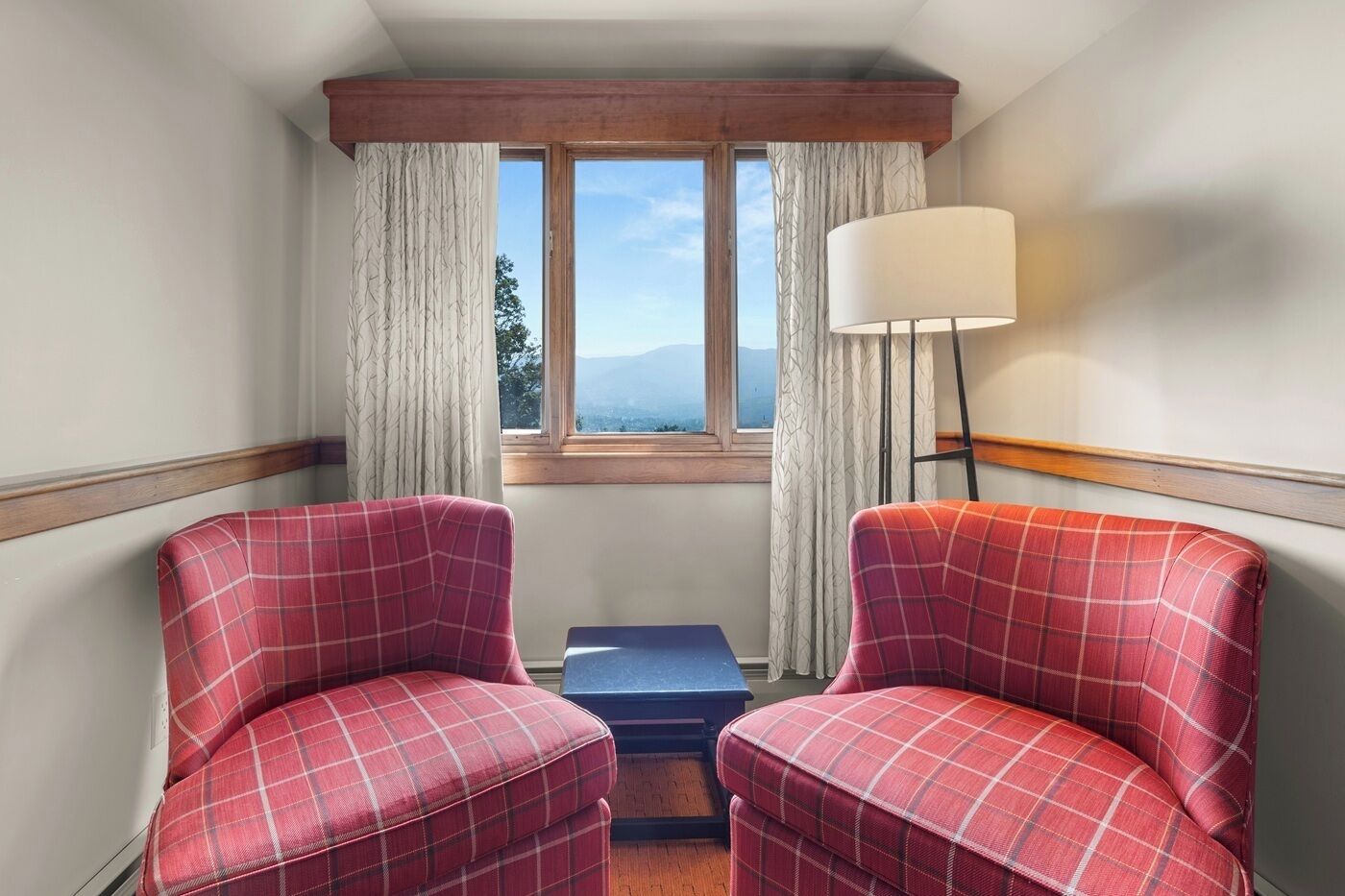 A corner room with two plaid chairs, a small table, a lamp, and a window with curtains.
