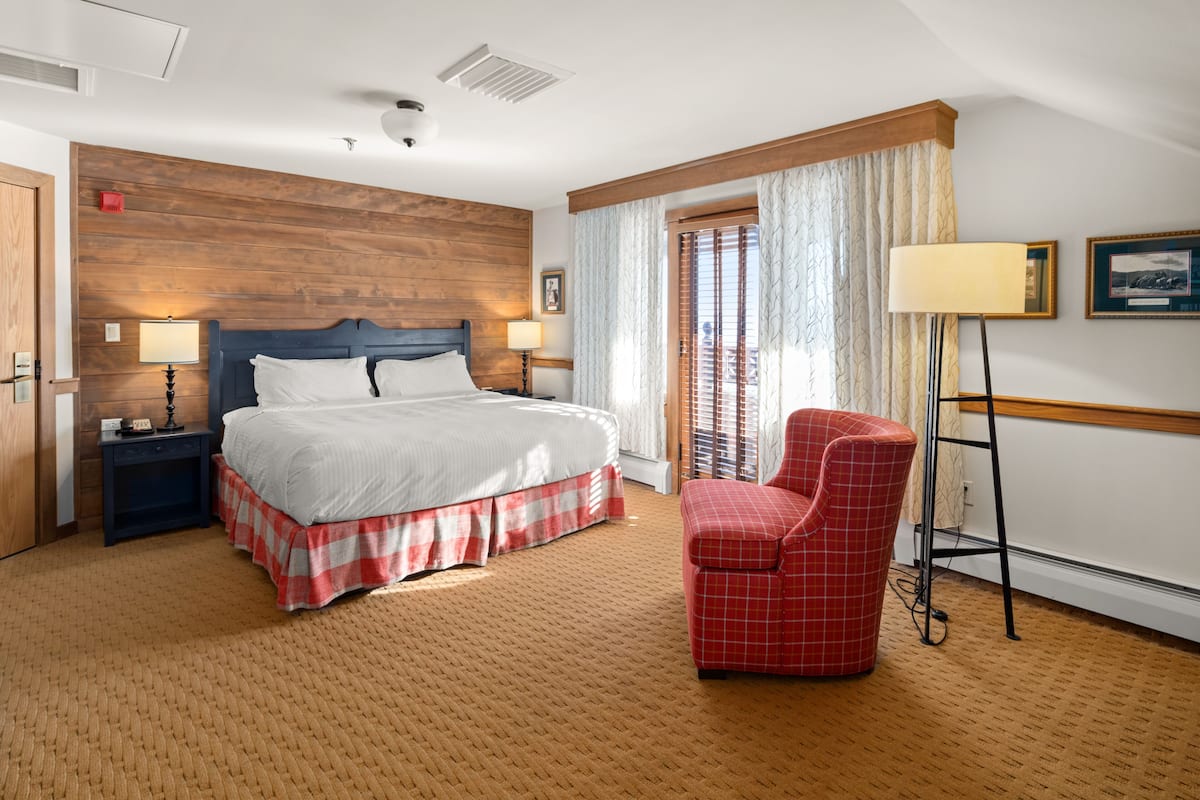 Una habitación de hotel con una cama grande, una silla de cuadros rojos, una lámpara de pie y una ventana con cortinas.