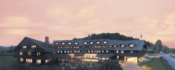 Um hotel grande, com vários andares e um pátio central imponente, rodeado por vegetação exuberante e montanhas ao fundo.