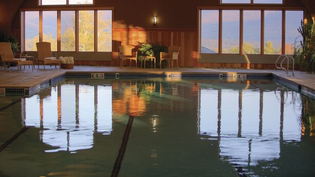 An indoor swimming pool with clear reflections, large windows, and a seating area with chairs and a table.