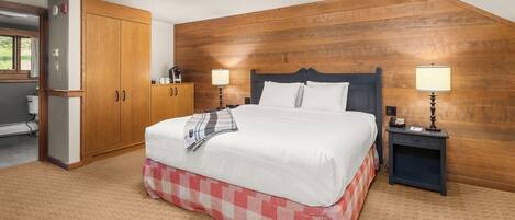 Une chambre à coucher comprenant un lit, une tête de lit en bois, deux tables de chevet avec des lampes, une armoire et un coin cuisine.
