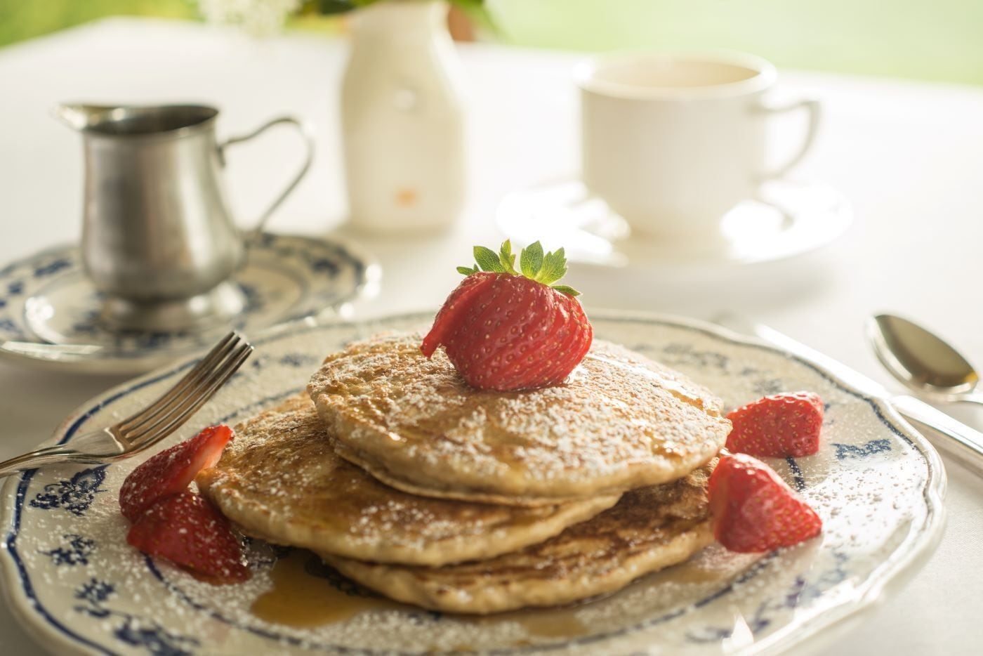 A plate of pancakes with strawberries and powdered sugar, a pitcher, a cup, and a spoon on a table.