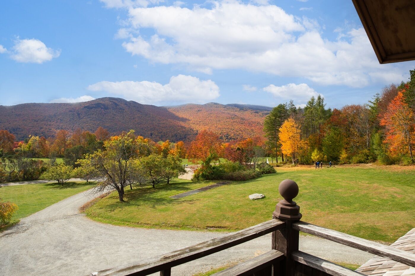 A scenic view from a balcony overlooking a grassy area with trees displaying autumn colors, a gravel path, and mountains in the background.